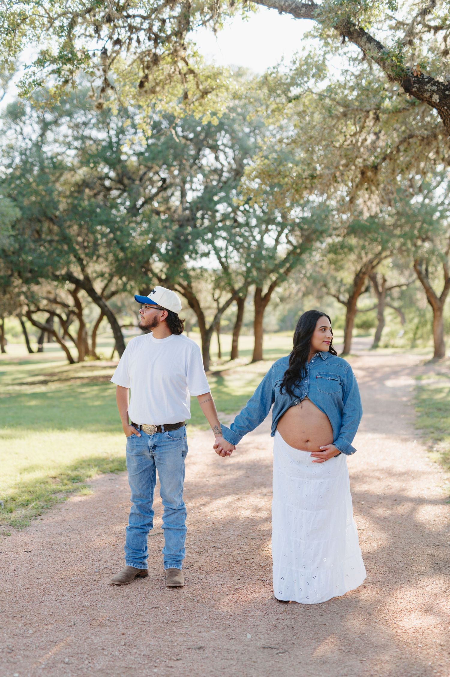 A couple holding hands on a tree-lined path as they pose for a maternity photoshoot in nature.