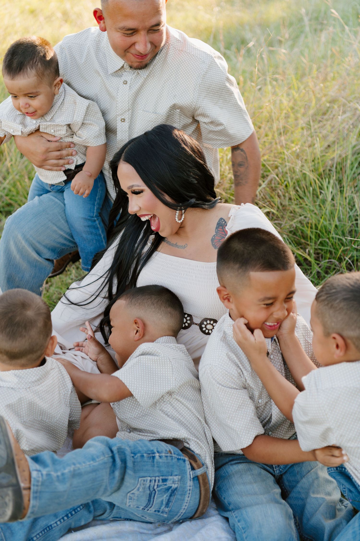 Family sitting together in tall grass field wearing coordinated white shirts and blue jeans during outdoor photo session.