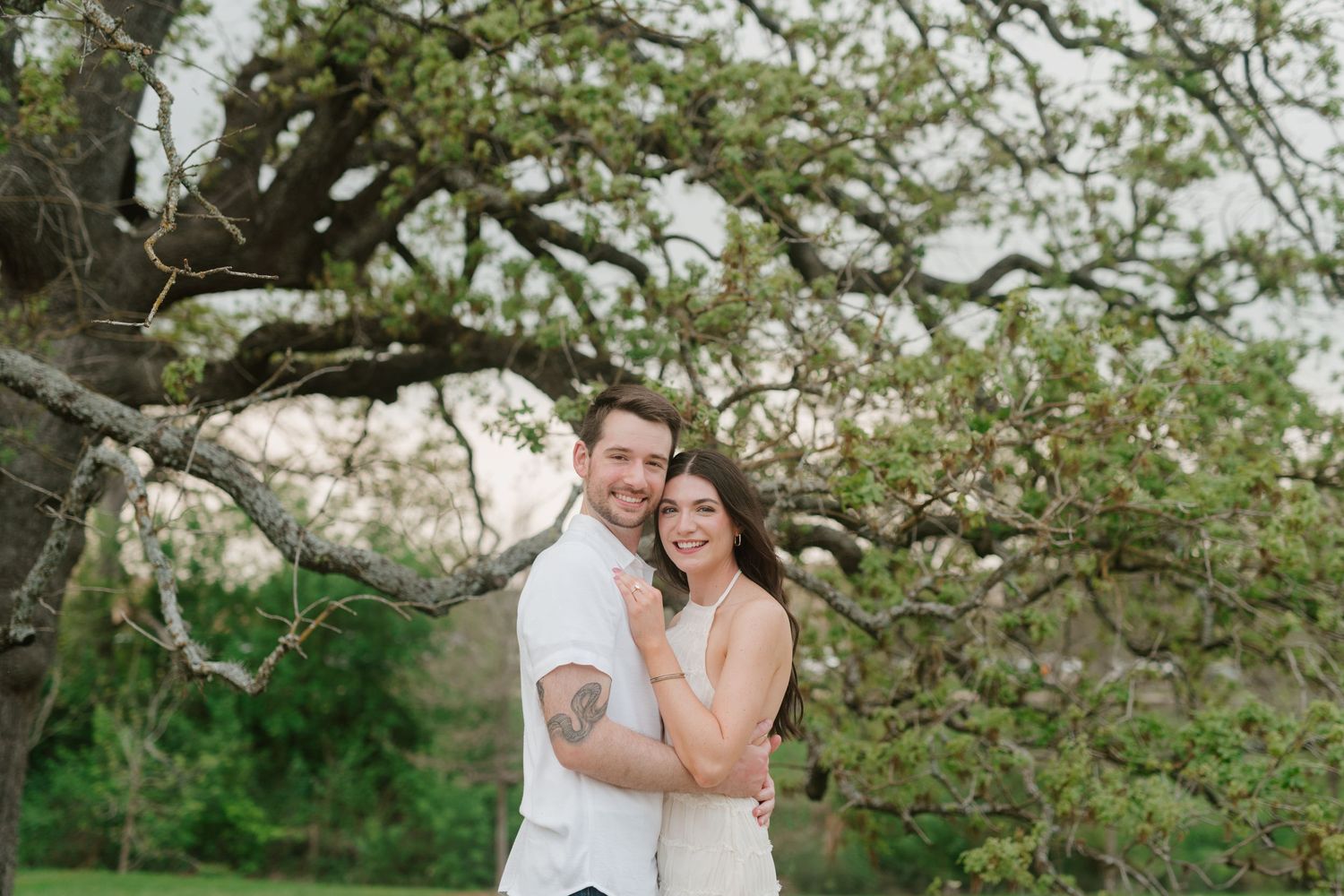 A couple embraces while standing beneath a sprawling tree branch in an outdoor setting during springtime.