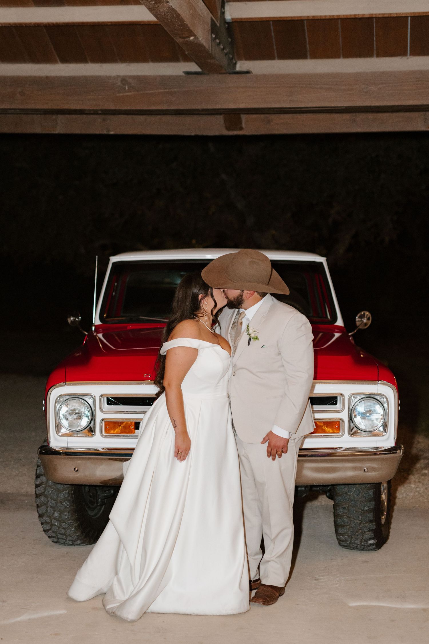 A newlywed couple shares a romantic moment in front of a classic red truck in a rustic barn setting.