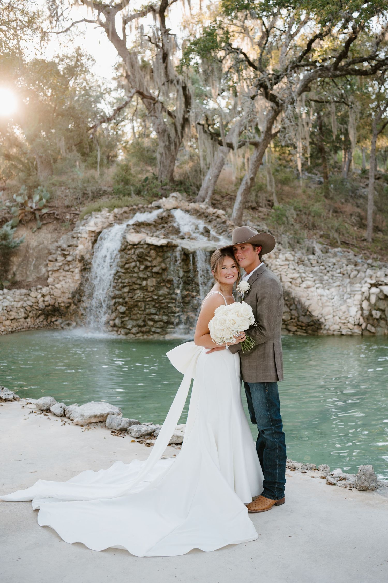 Newlyweds embrace by a turquoise pool with flowing white dress and rustic scenery.