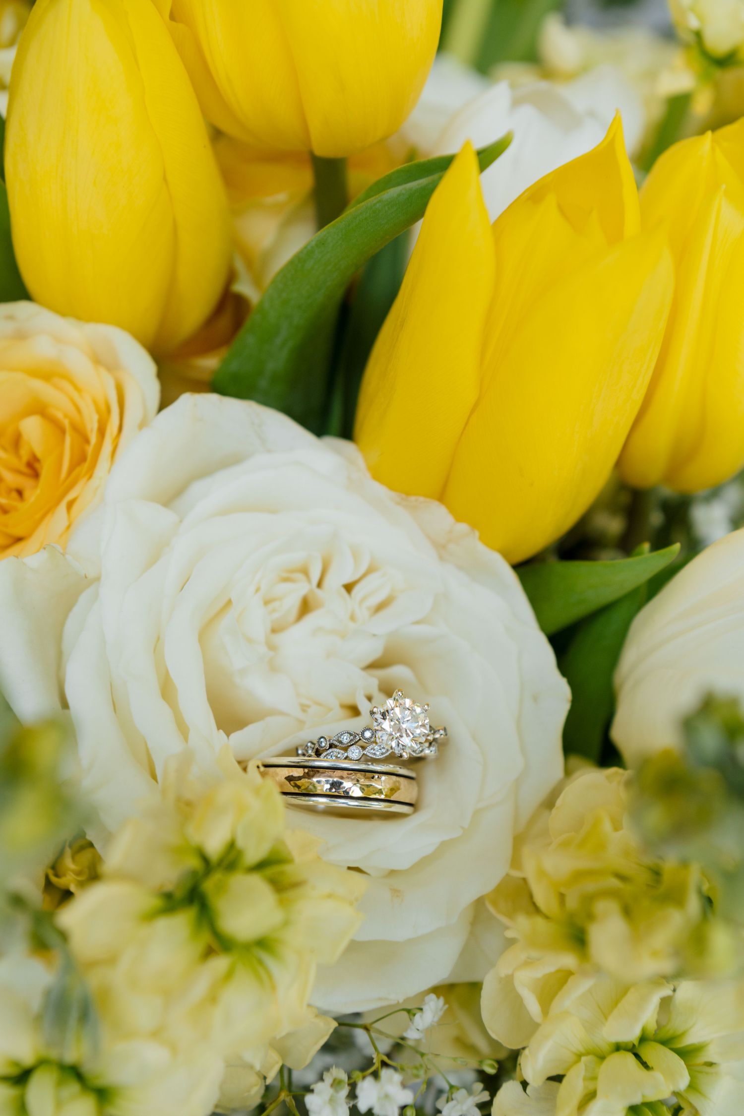 Gold wedding rings rest on white roses surrounded by yellow tulips in a bridal bouquet.