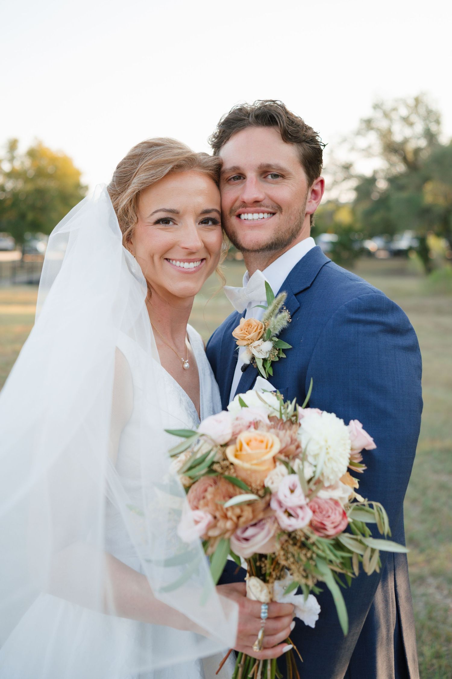 A bride and groom pose outdoors with a peach and white floral bouquet during their golden hour wedding portraits.
