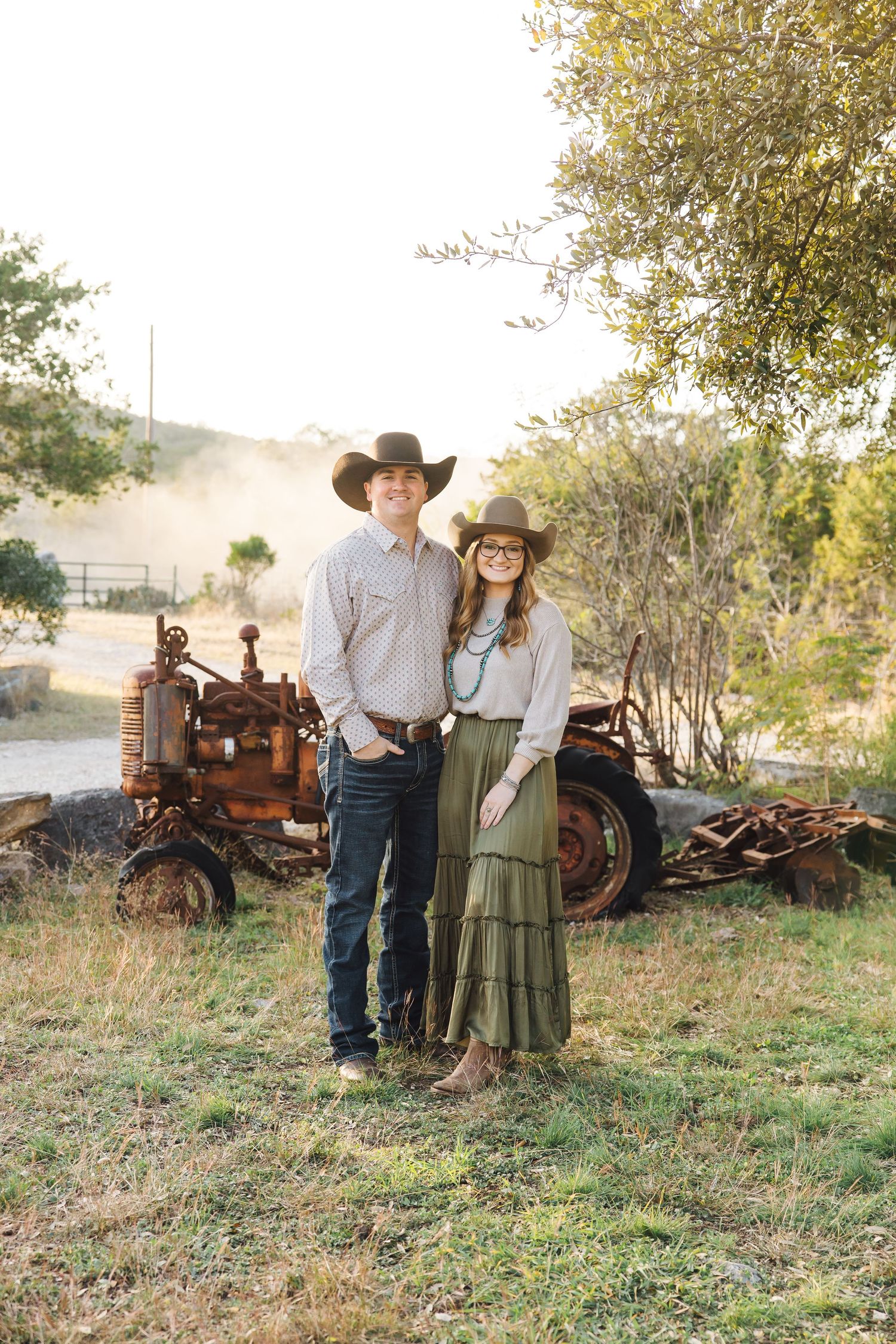 A couple poses near a vintage rusty tractor in a sunlit rural field.