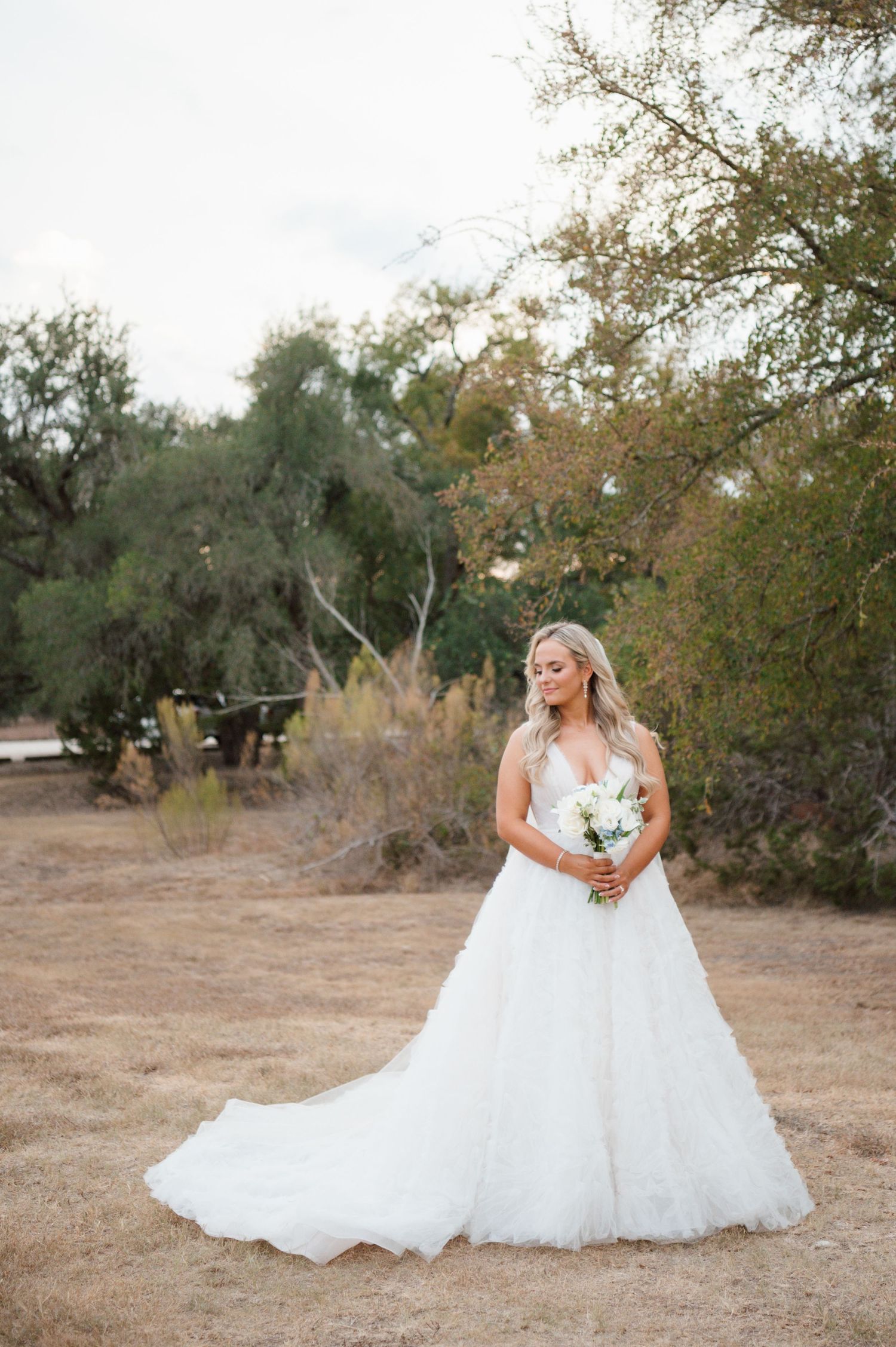 A bride standing in a field wearing a large, flowing wedding dress and holding a bouquet of flowers.