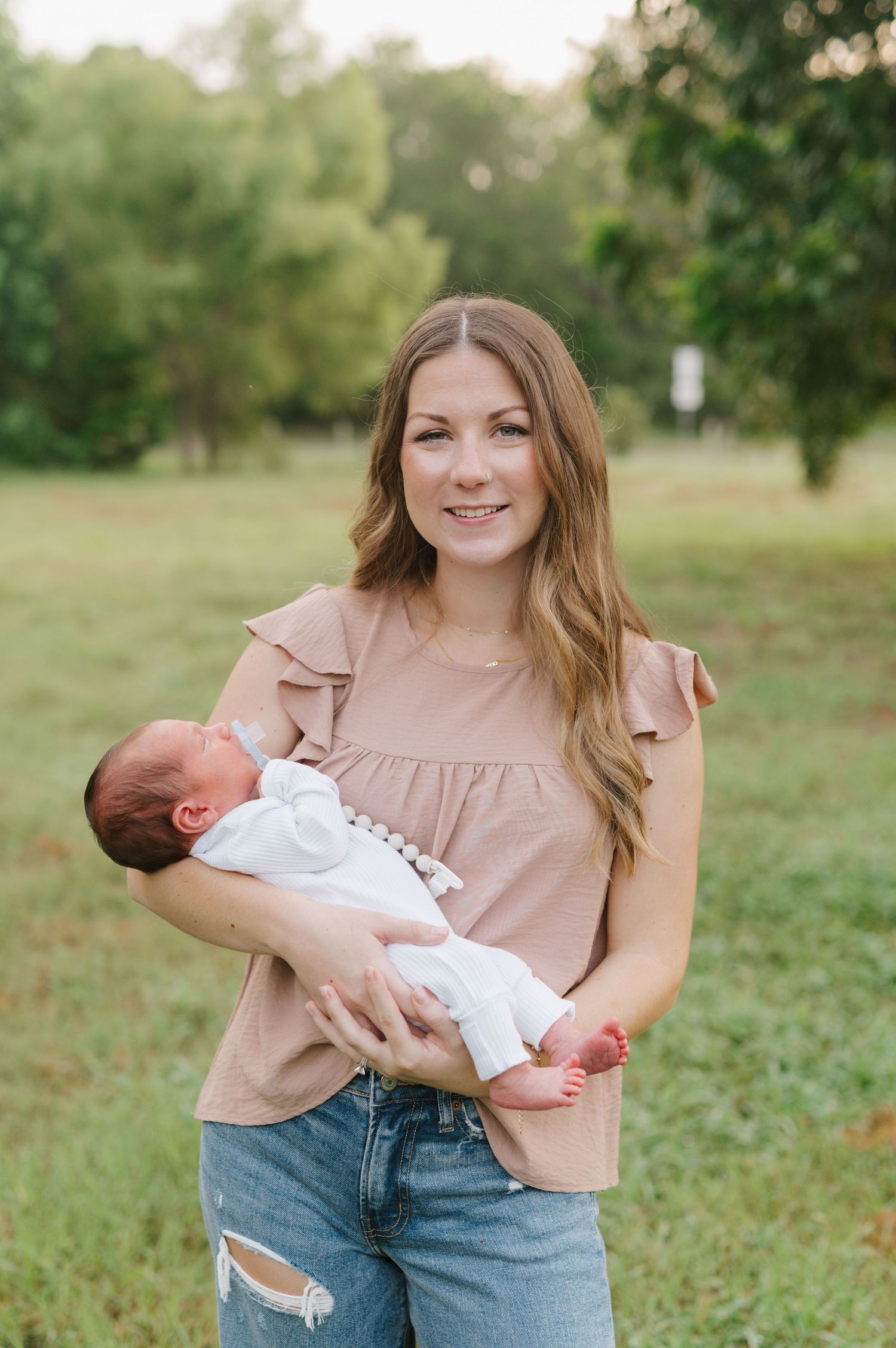 A woman in a mauve blouse and ripped jeans holds a sleeping newborn baby outdoors in a grassy park setting.