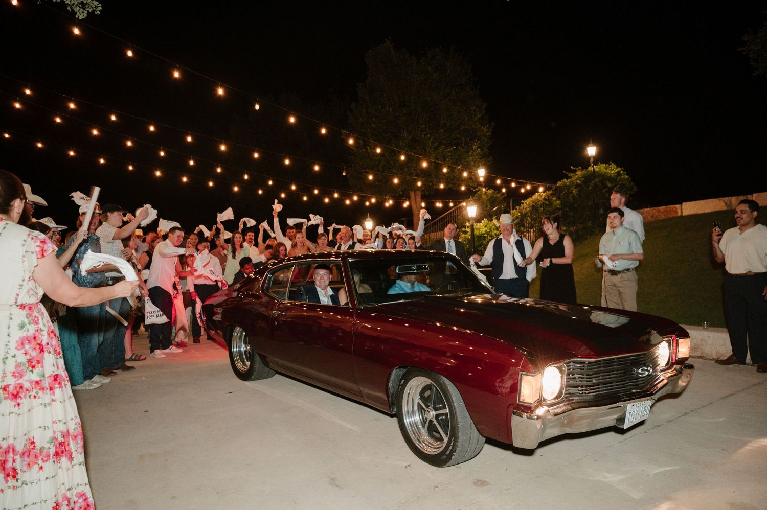 Classic burgundy vintage car departing under string lights with crowd waving sparklers.