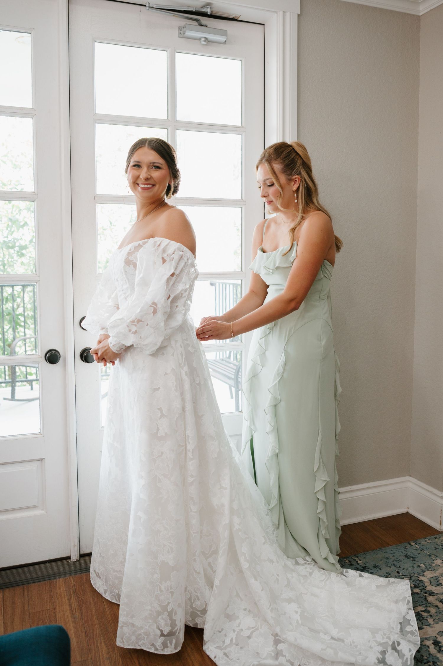 Bridesmaids in mint green dresses help adjust a flowing white lace wedding gown during pre-ceremony preparations.