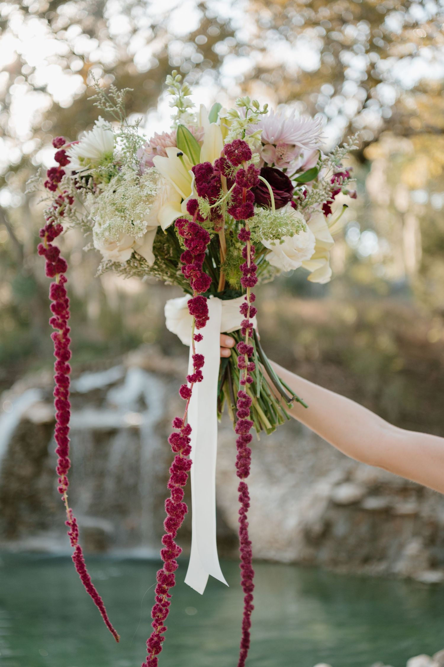 Wedding bouquet with cascading deep red amaranthus and white flowers against a waterfront backdrop.