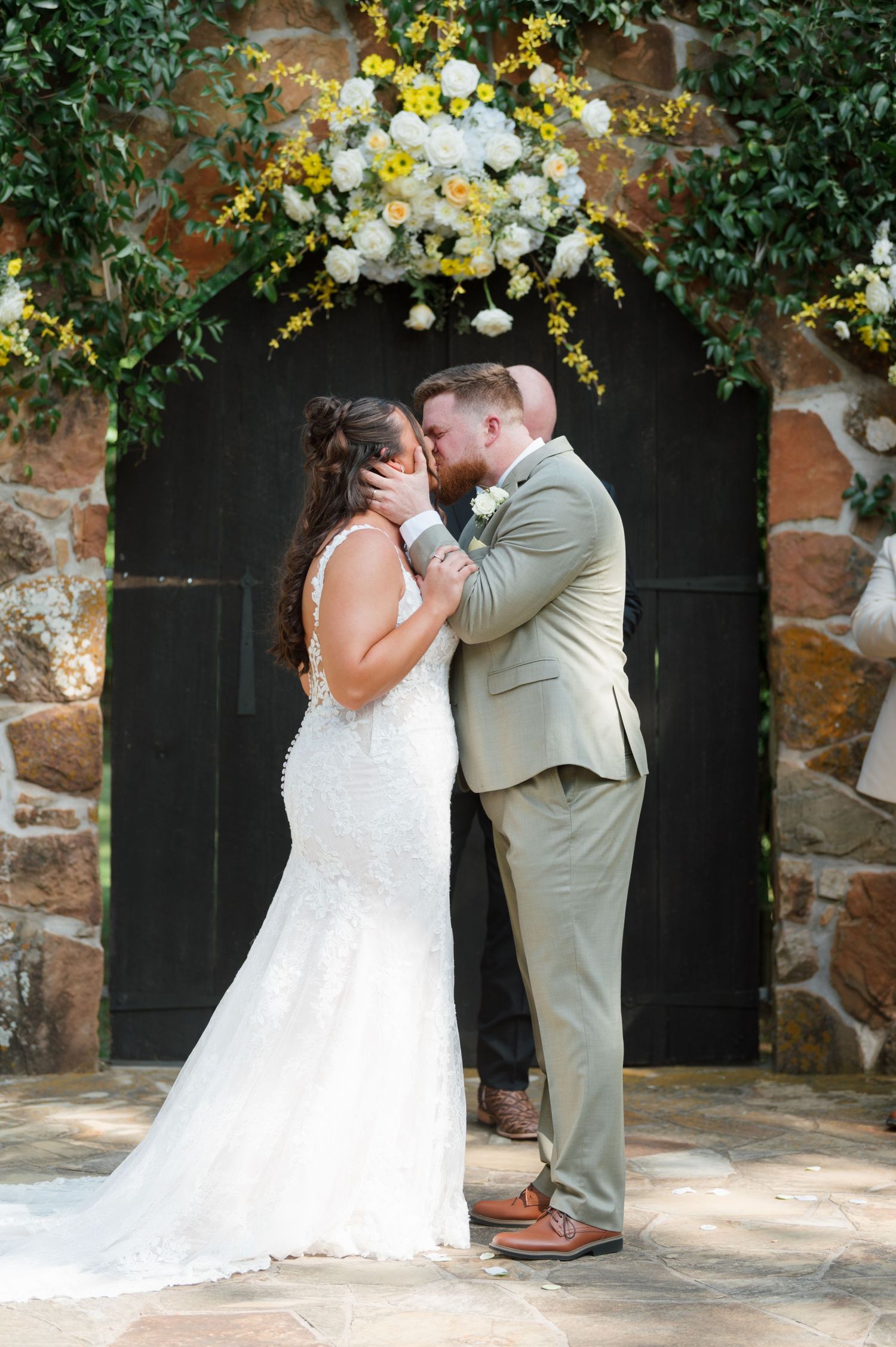 Bride and groom share their first kiss at an outdoor ceremony adorned with white and yellow floral arch.