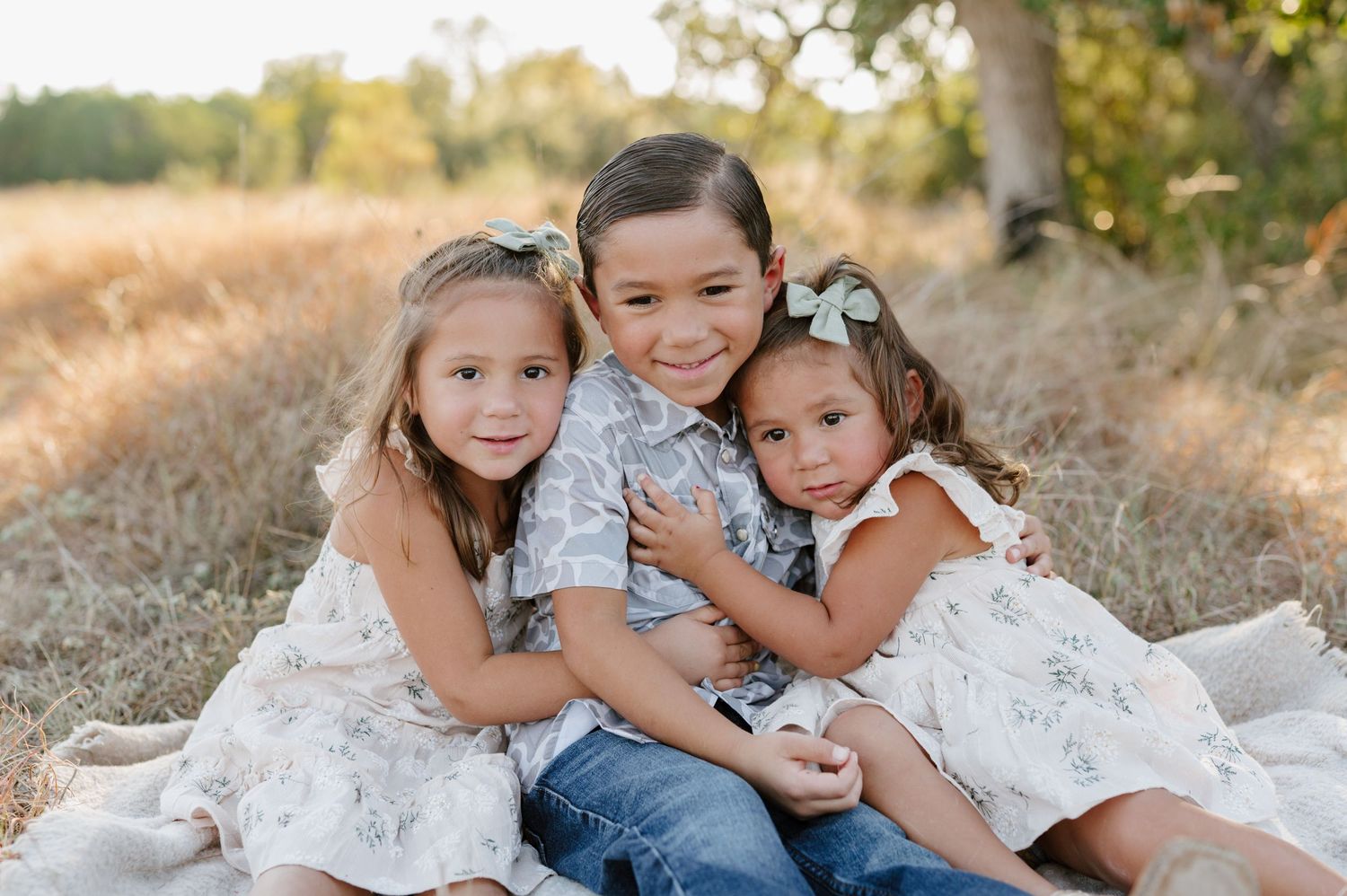 A group of children wearing white and denim sit together on dry grass during a sunny outdoor photoshoot.