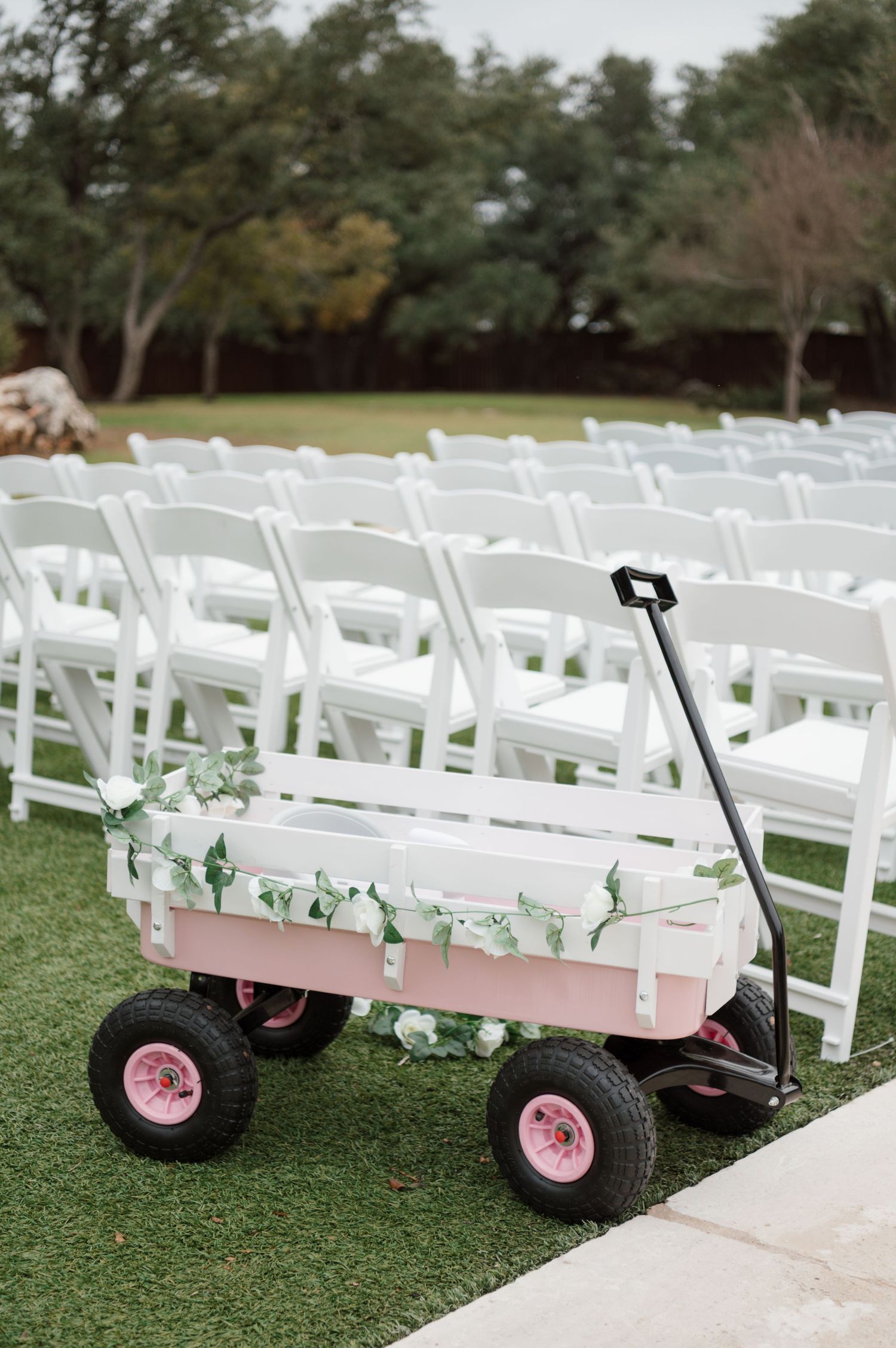 Pink wagon with white decorations sits on grass near rows of white folding chairs at an outdoor wedding setup.