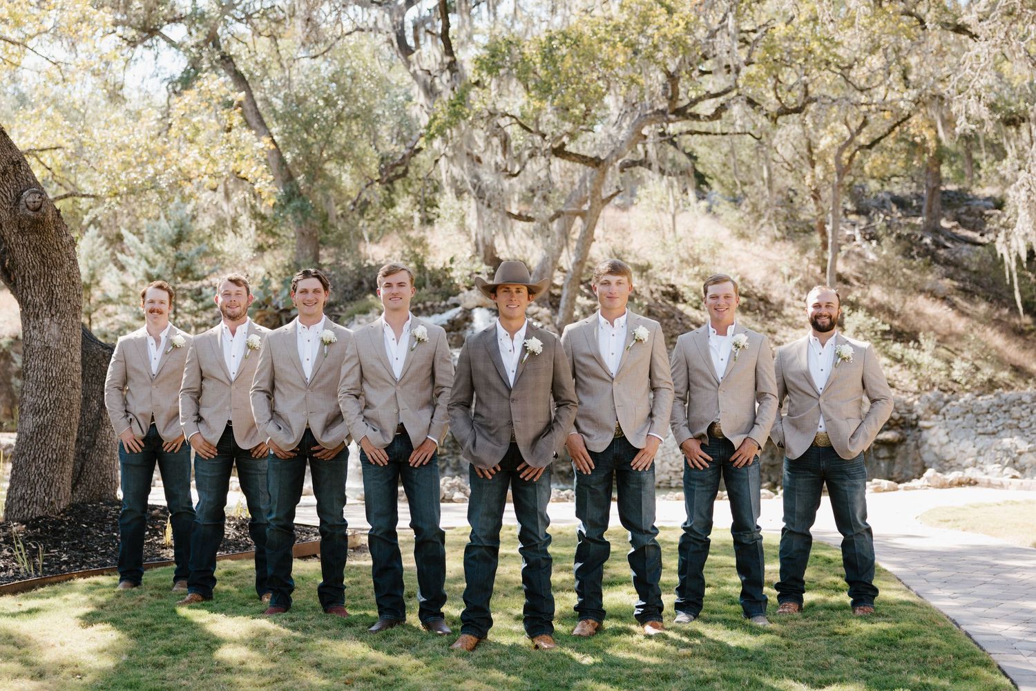 Groomsmen in light gray suits stand in a row outdoors under trees on a sunny wedding day.