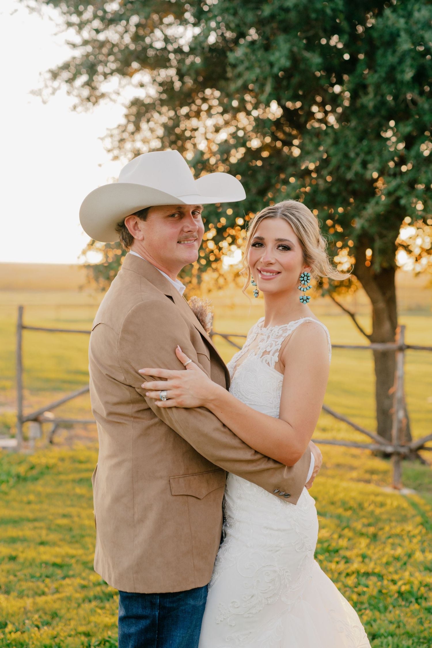 A romantic outdoor golden hour portrait of a couple in western wedding attire standing in front of a rustic fence.