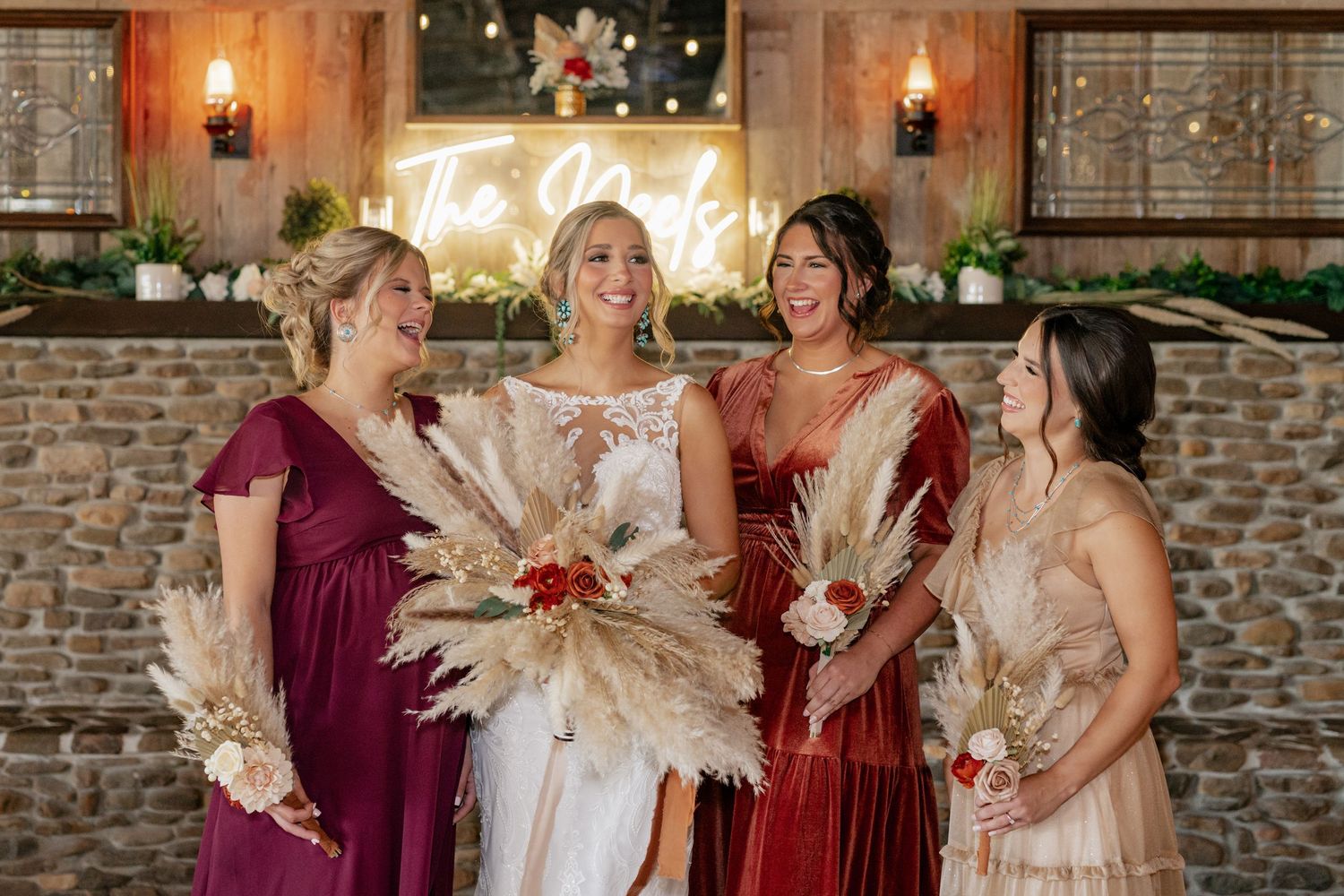 Wedding party in burgundy and blush dresses holding pampas grass bouquets against a stone wall.