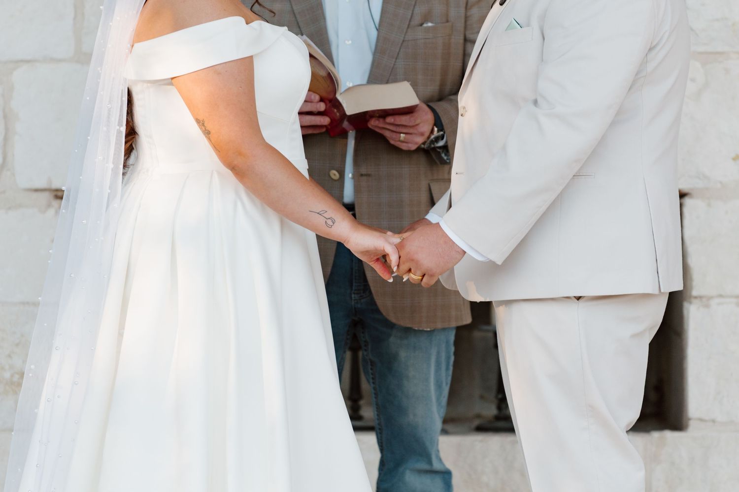 A couple holds hands during an intimate wedding ceremony, with the bride wearing a white off-shoulder dress.