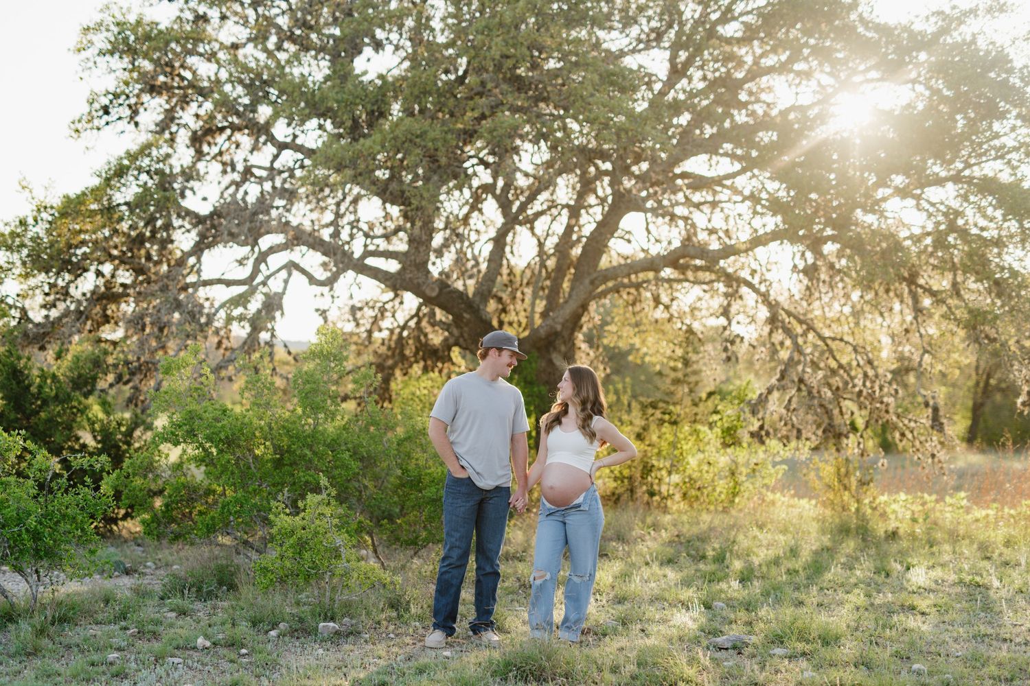 A couple stands together against a sunlit oak tree backdrop during golden hour in a natural outdoor setting.