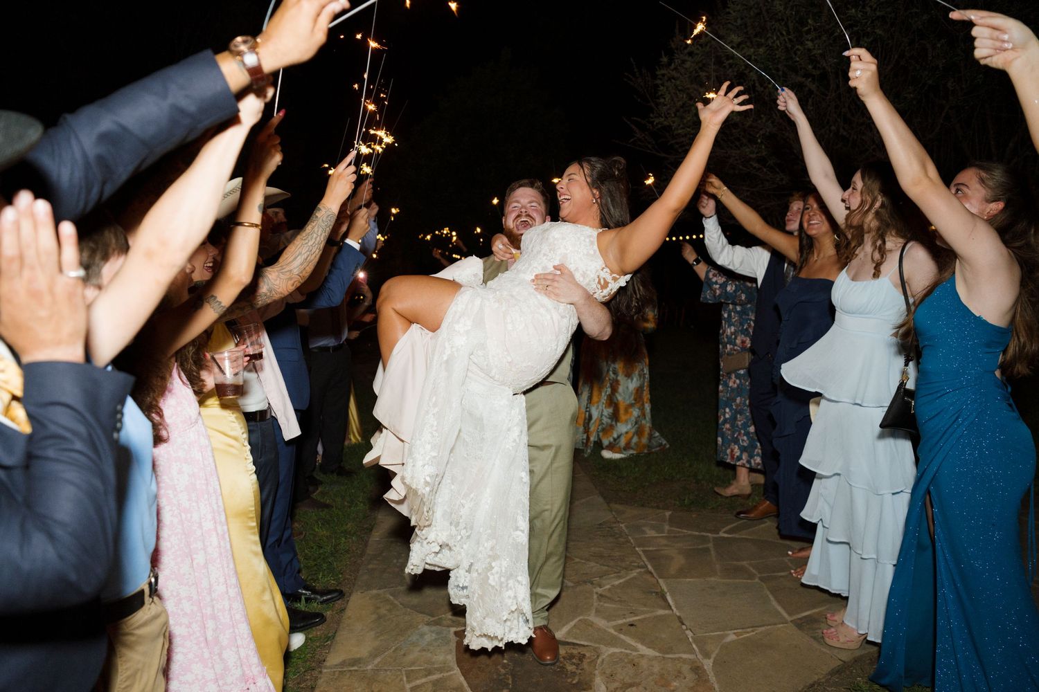 Groom lifts bride in wedding dress during sparkler send-off exit surrounded by cheering guests at night.