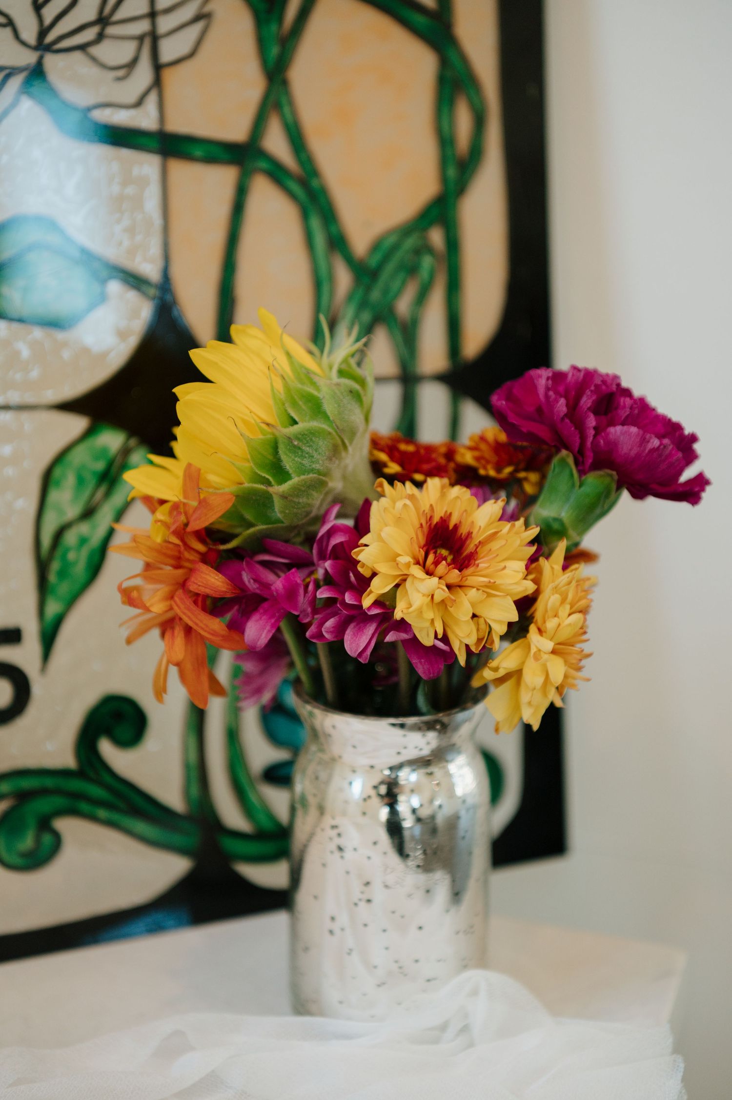 Colorful yellow, orange and purple flowers arranged in a mercury glass vase against a light wall.