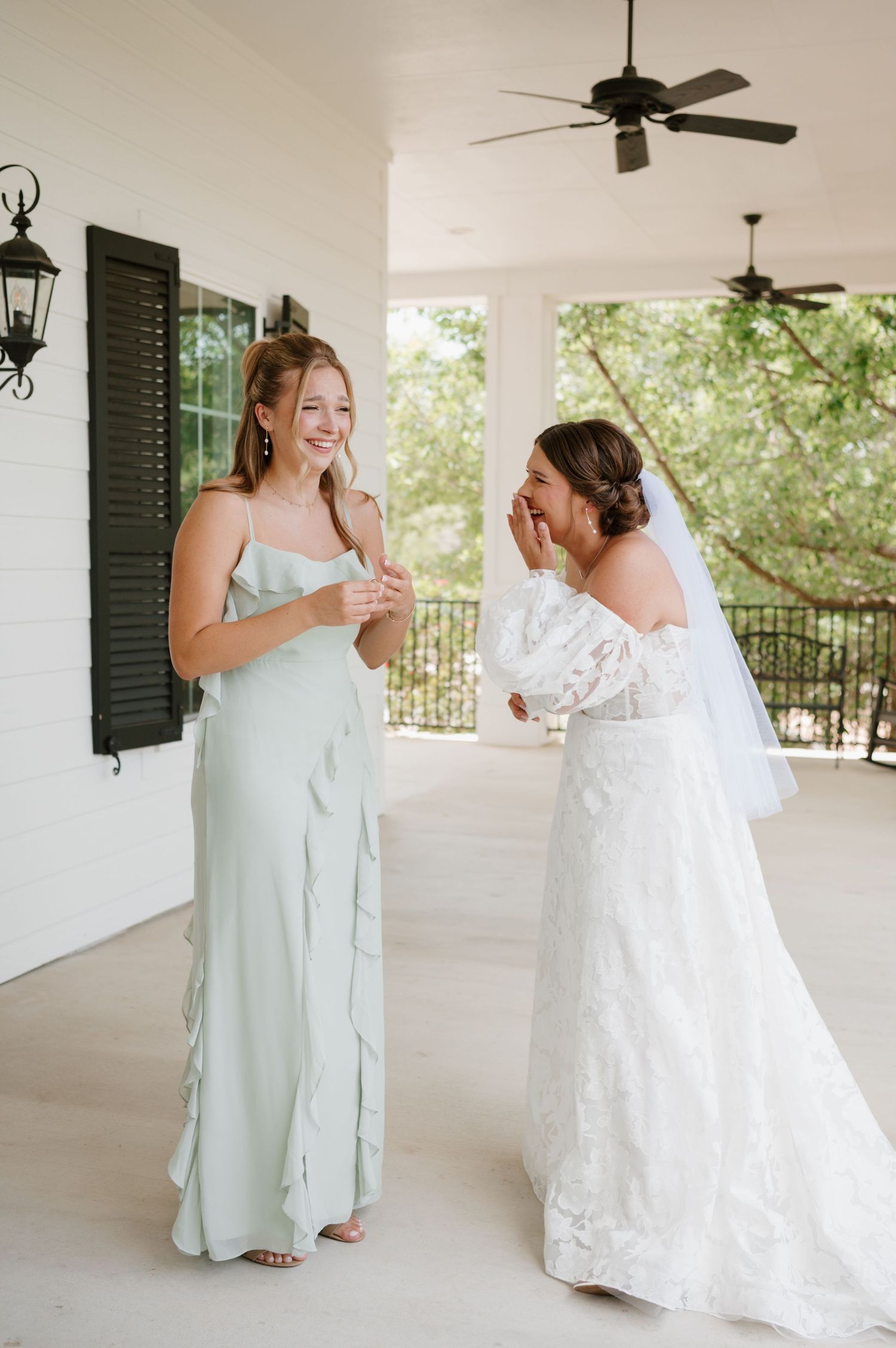 Two women share a joyful moment on a white porch, one in a white wedding gown and one in mint dress.