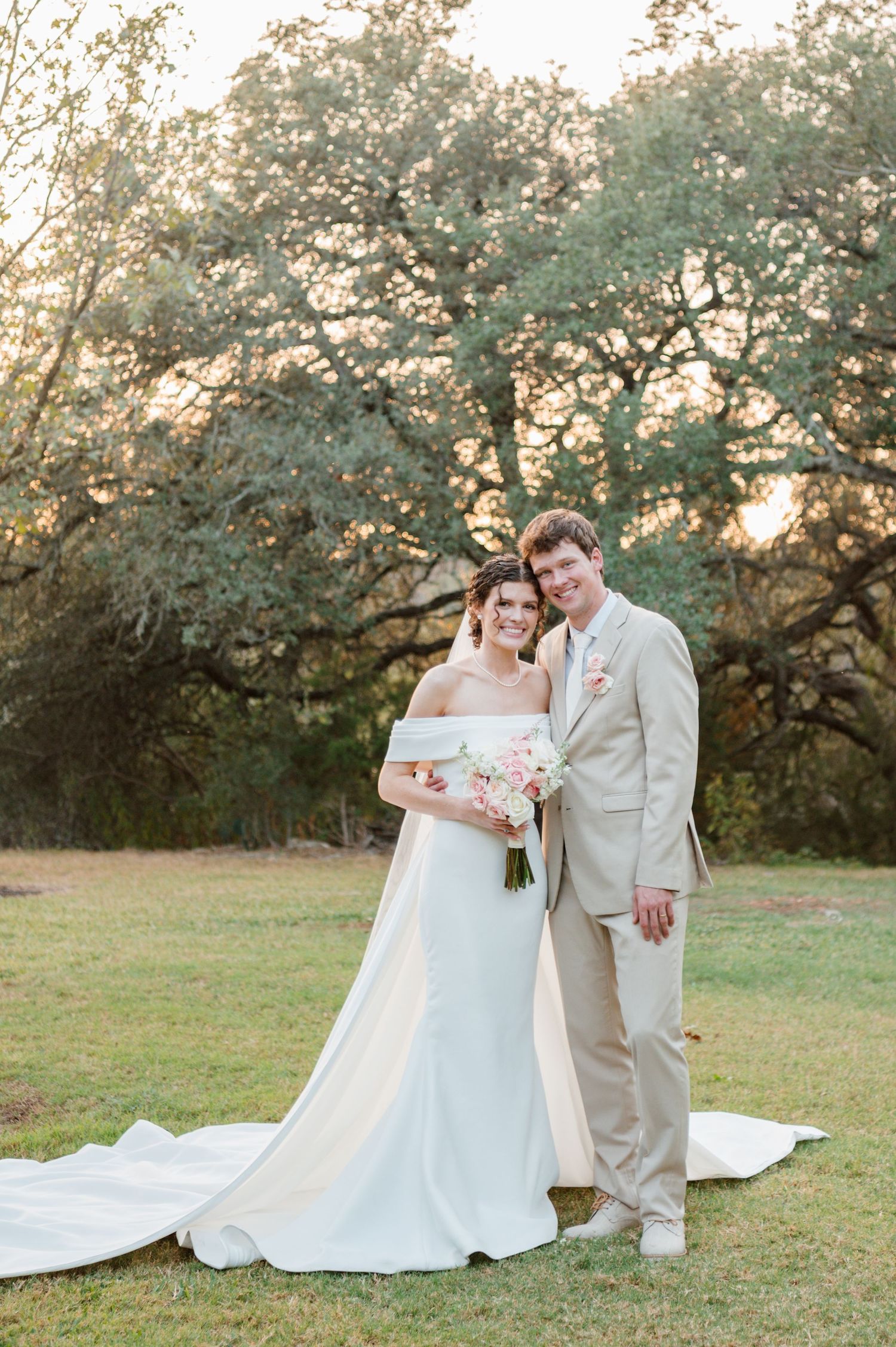 Couple in wedding attire poses on lawn with blooming trees in background during golden hour.