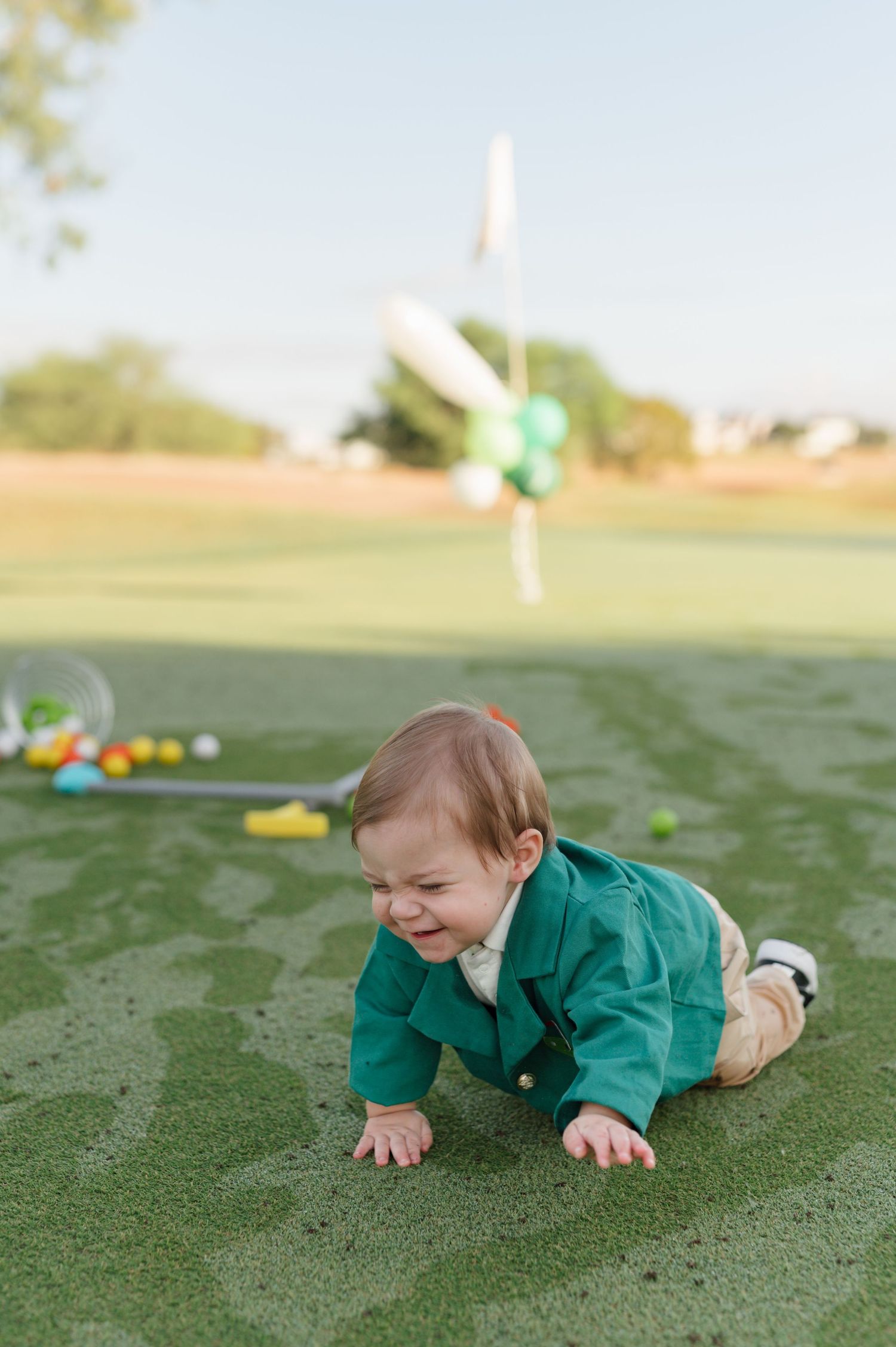 A small child in a green outfit crawls and plays on a grassy field during a sunny day.