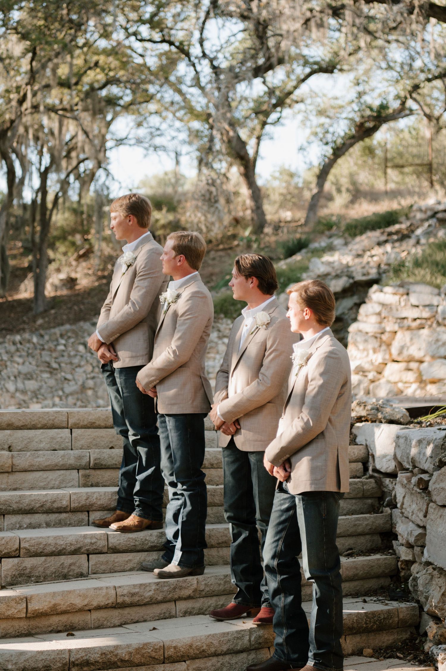 Groomsmen in tan jackets and jeans sitting casually on stone steps.