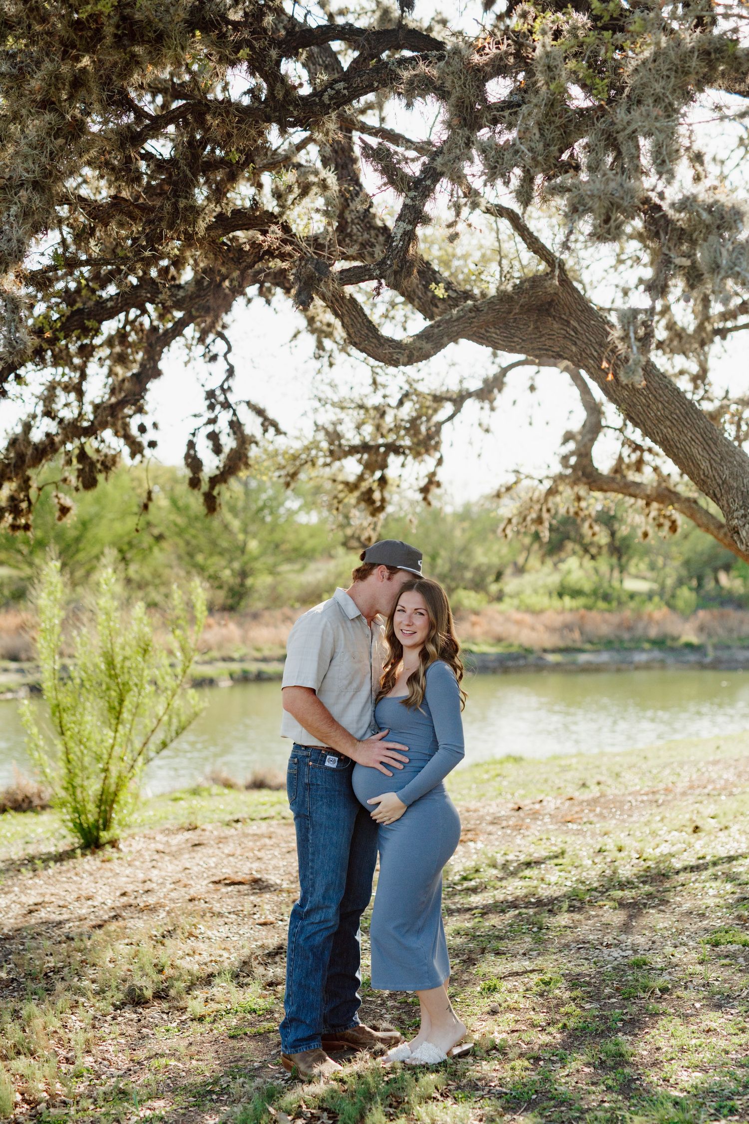 Couple embraces under large tree branch by tranquil lake during golden hour maternity photoshoot.