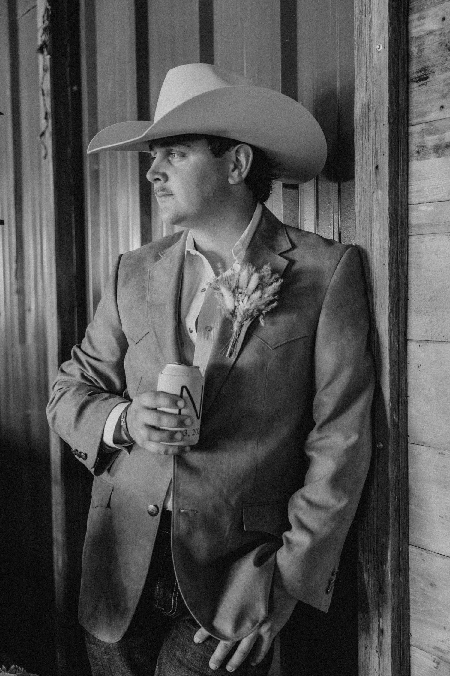 A western-styled groom in a cowboy hat and suit holding a drink at a rustic venue.