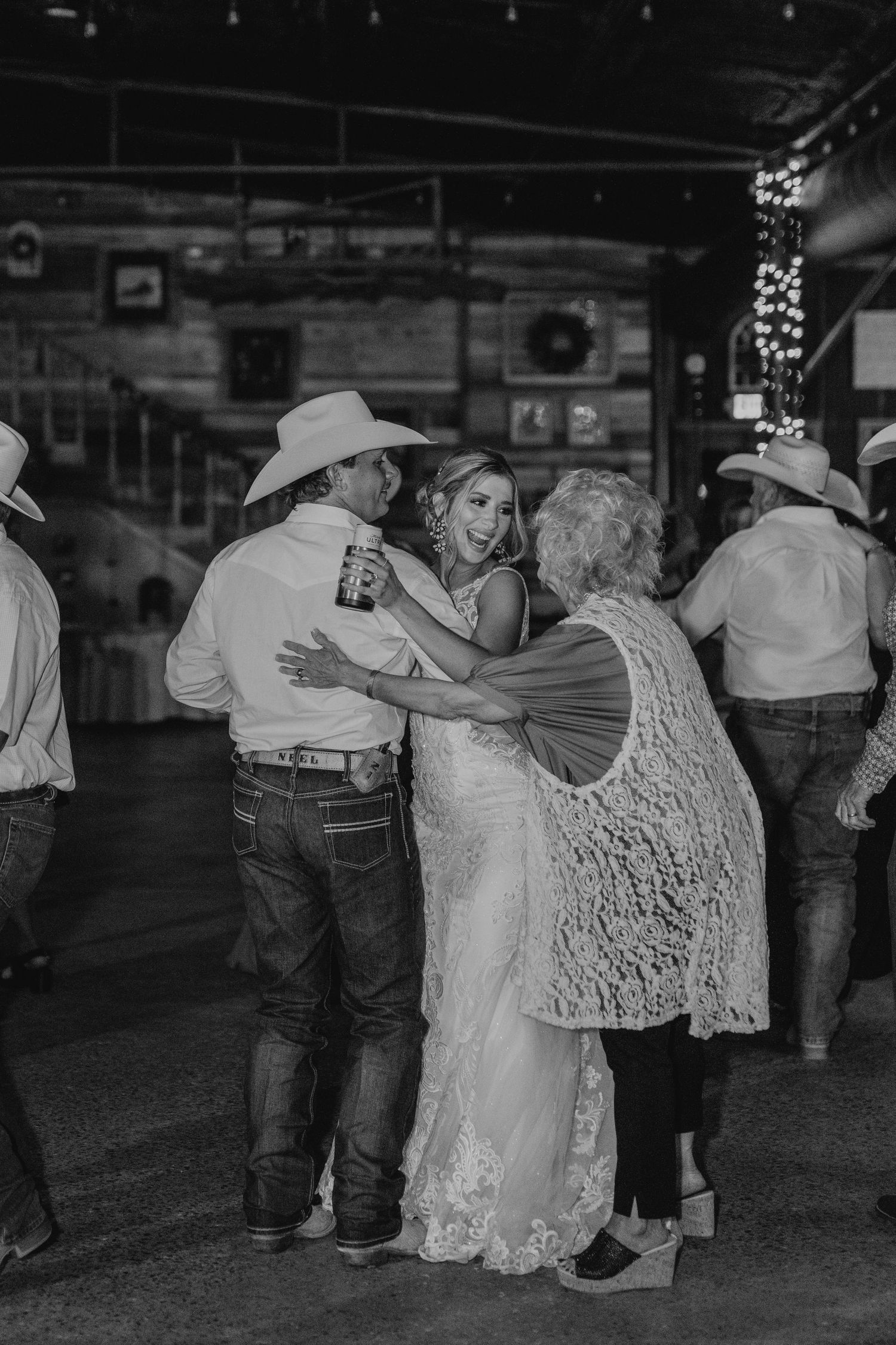 A black and white photograph captures wedding guests dancing and mingling at a rustic venue with string lights overhead.