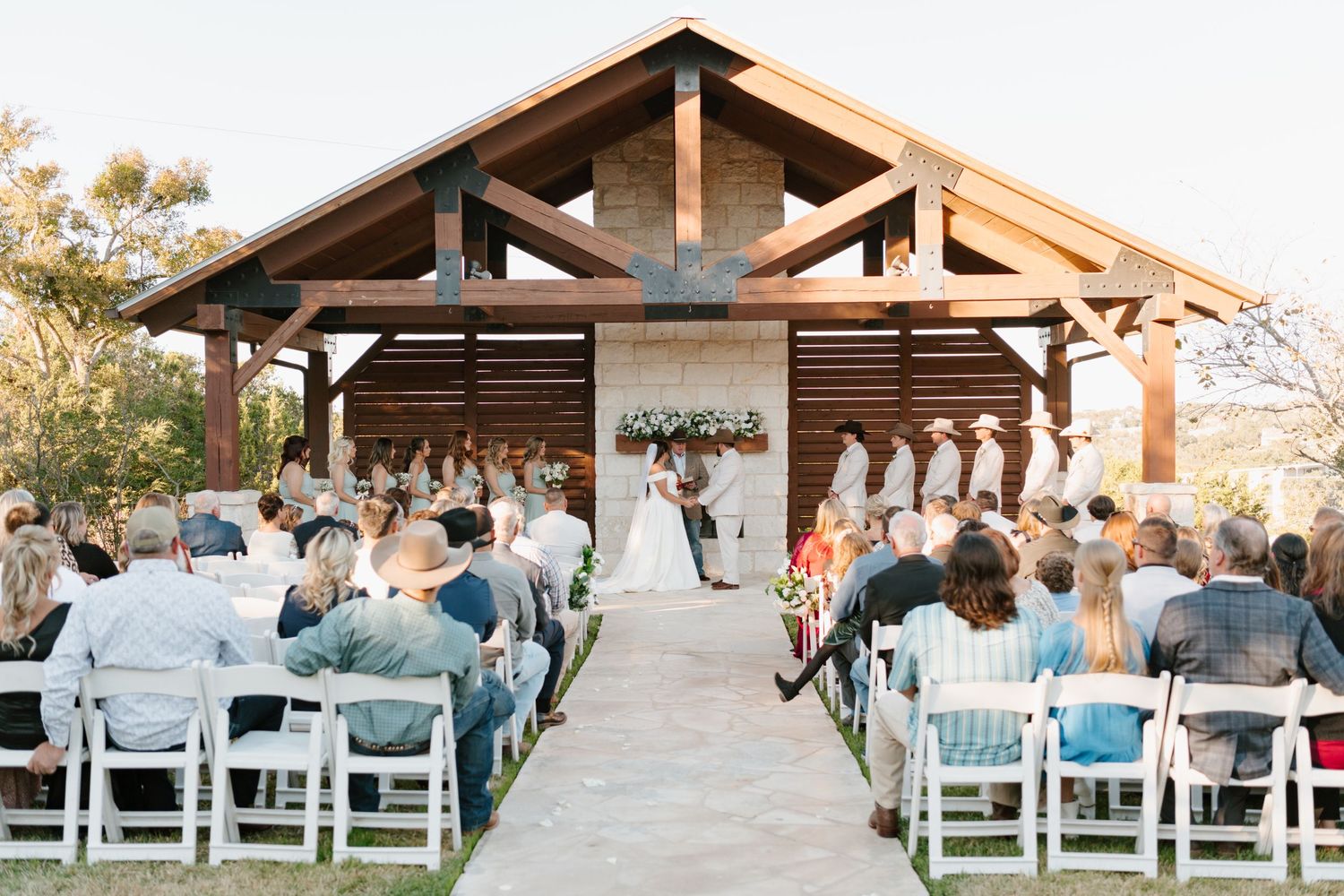Outdoor wedding ceremony under wooden pavilion with guests seated in white chairs facing floral-decorated altar.