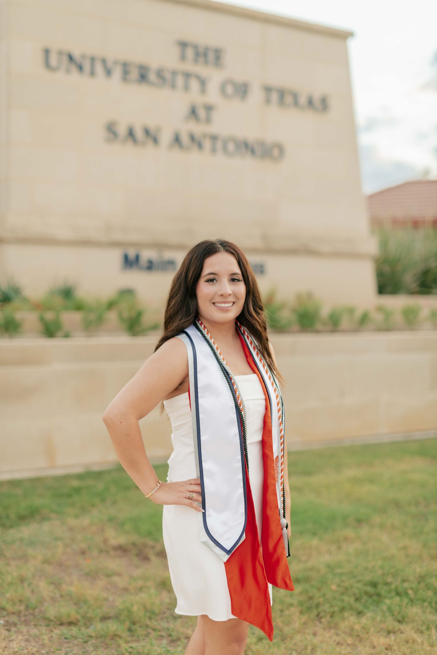 Graduate in white dress and red stole poses in front of University of Texas at San Antonio sign.