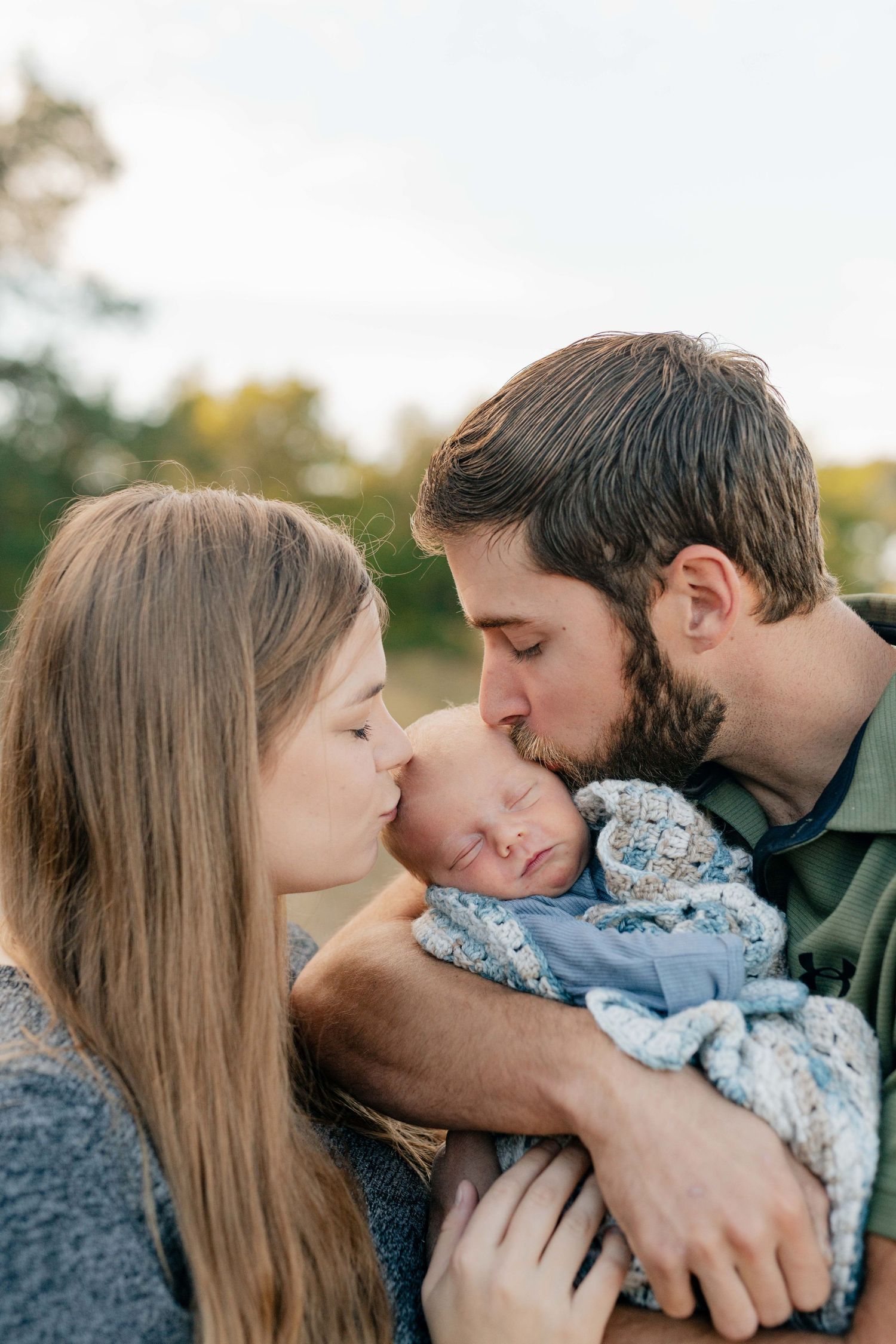 Intimate family moment sharing kisses with newborn against natural outdoor backdrop.