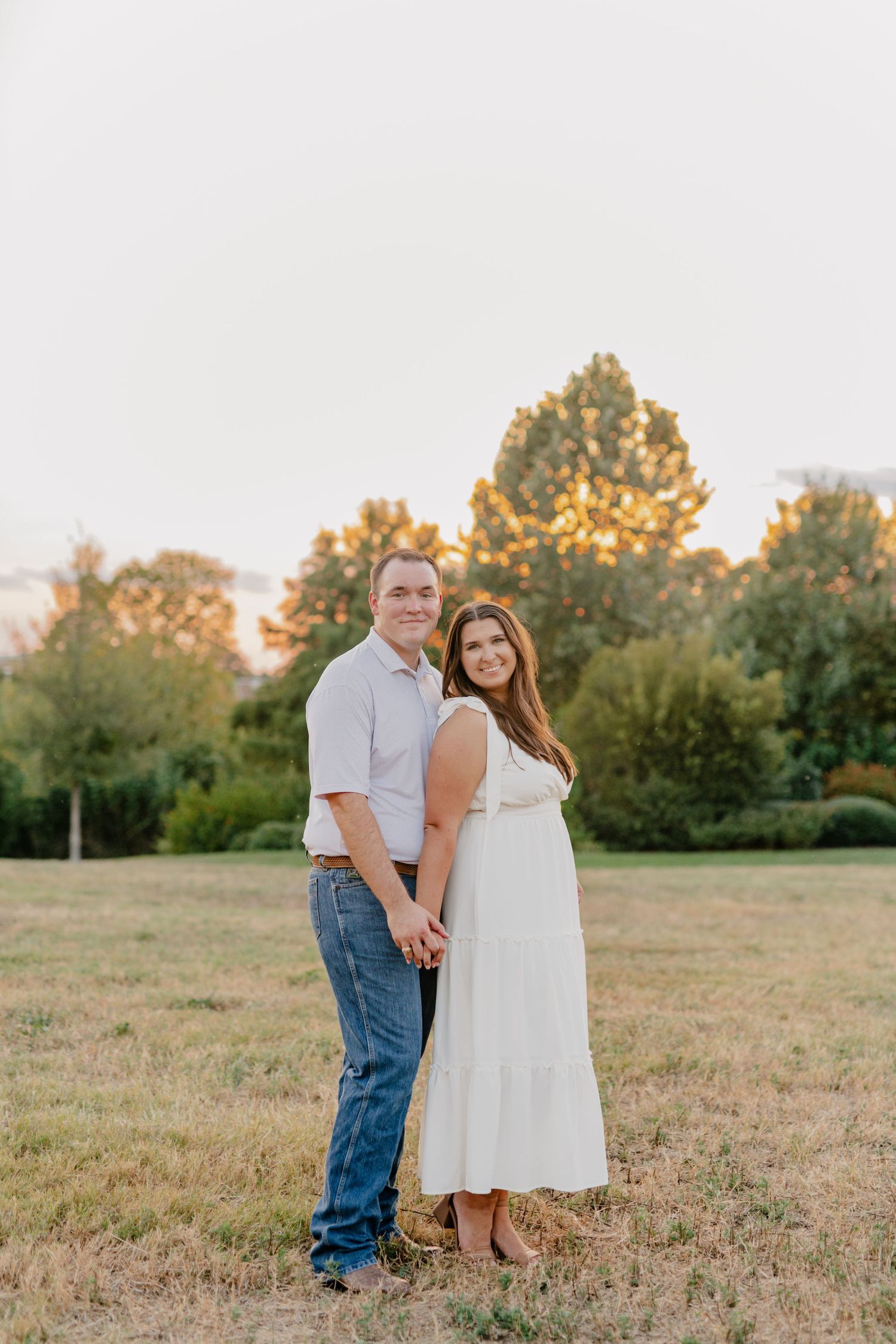 A couple poses together in a grassy field at sunset with trees silhouetted in the background.