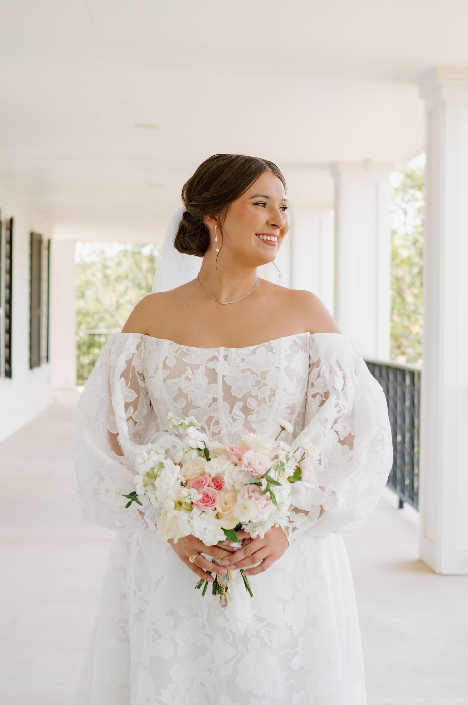 Elegant bride in off-shoulder lace gown holds pink and white bouquet on white porch.