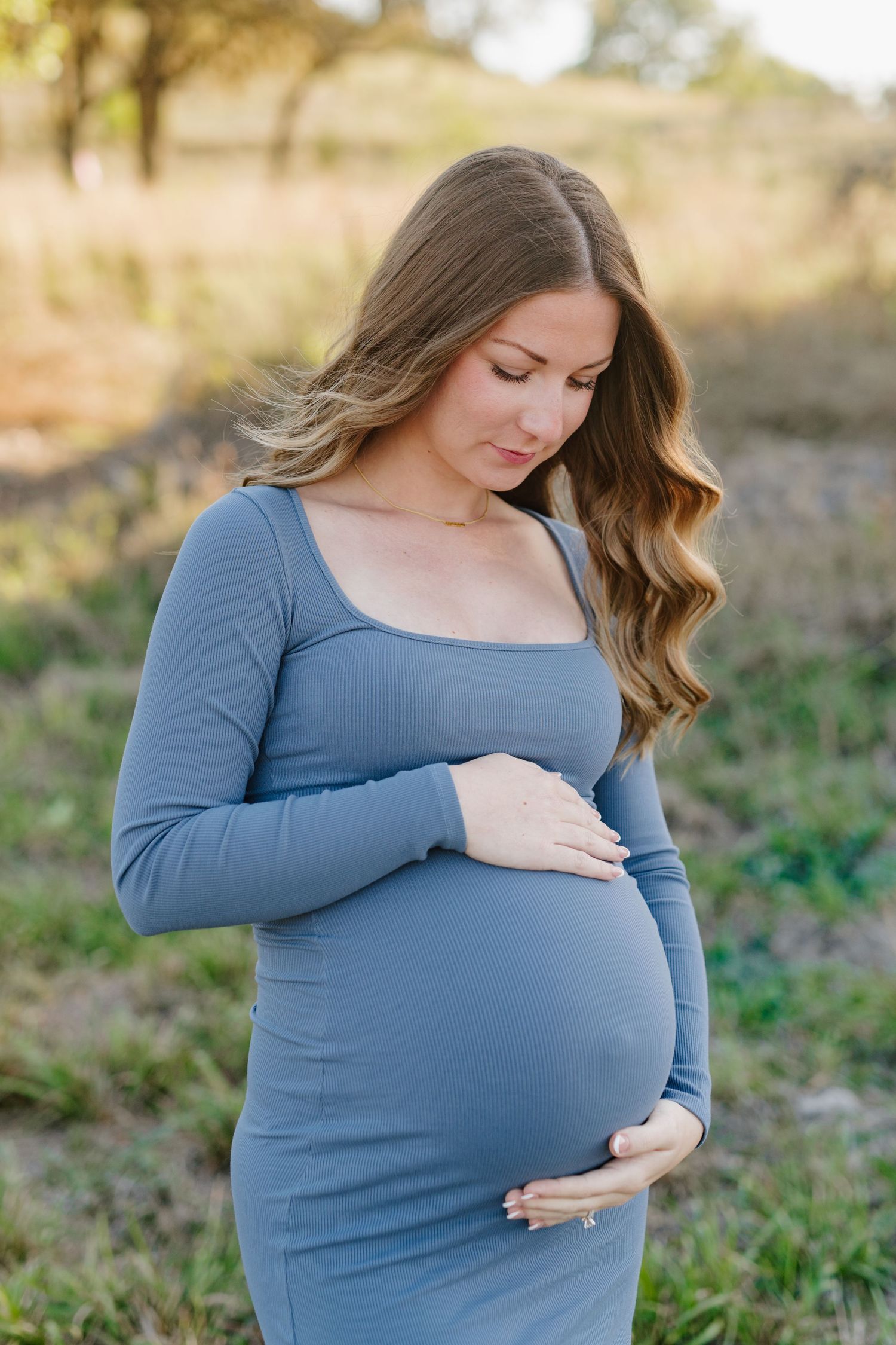 An expectant mother in a fitted blue dress gently touches her baby bump in an outdoor setting.