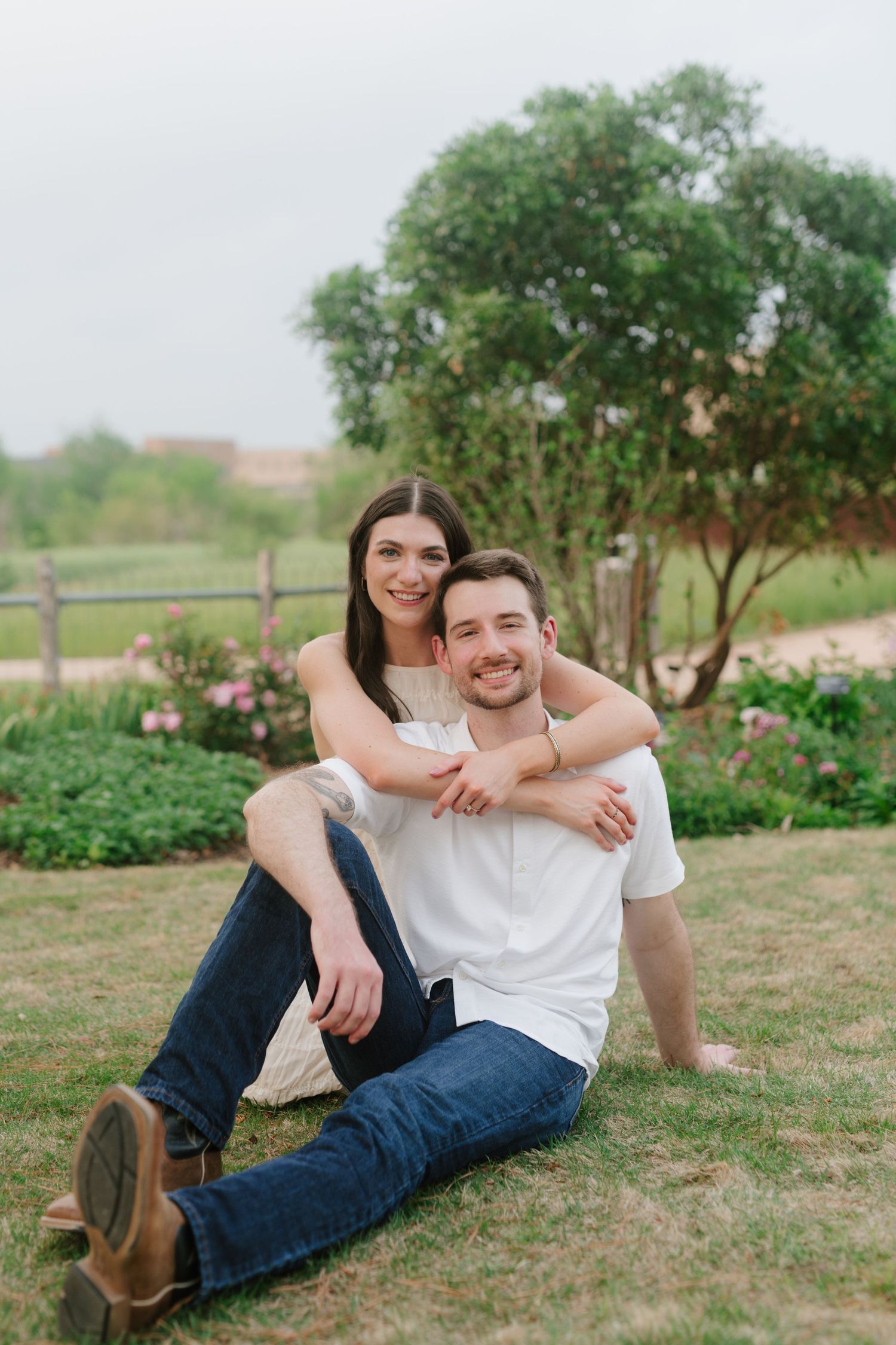 Happy couple sits together on grassy lawn with flowering garden and rustic fence in background.