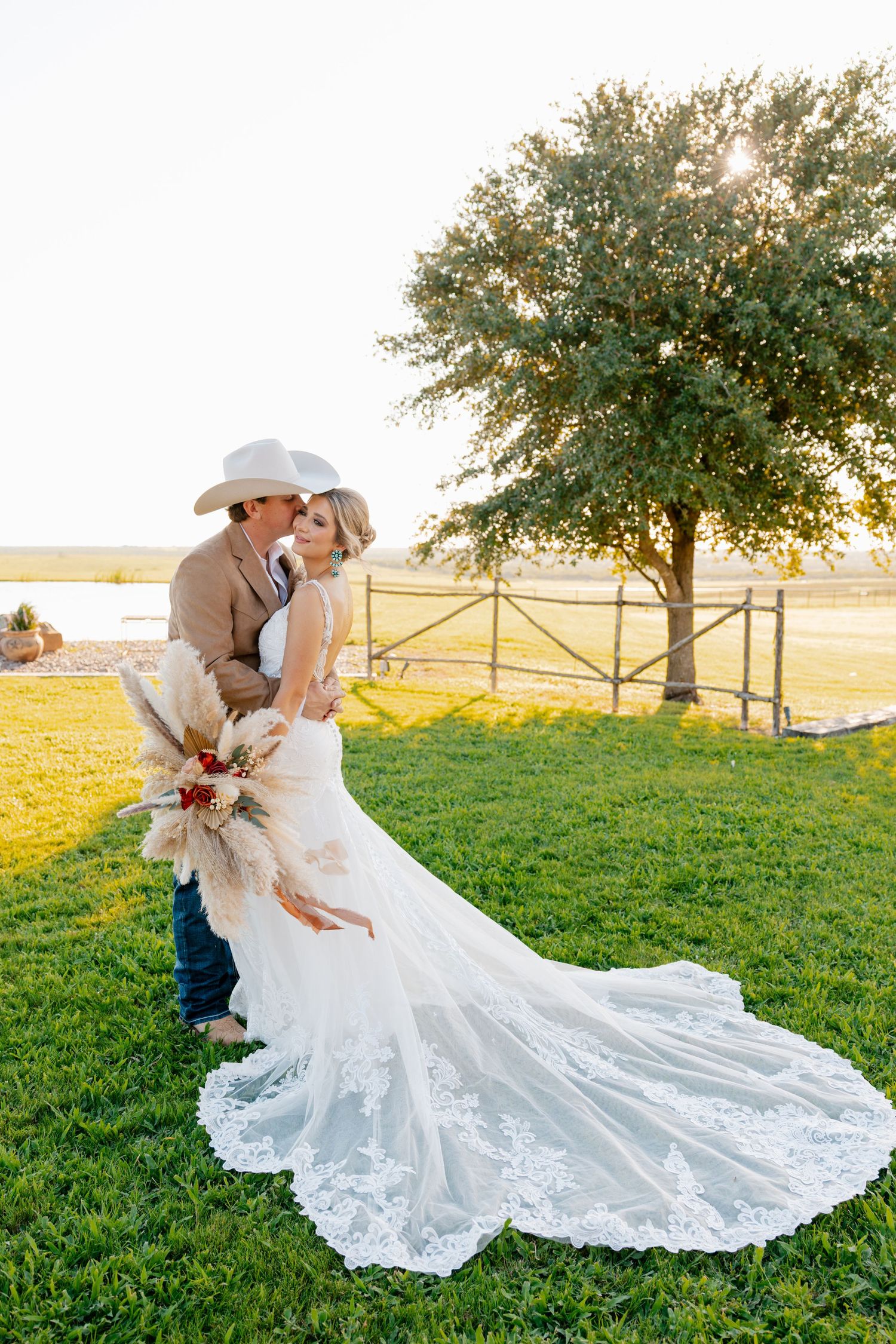 Romantic sunset wedding photo of couple embracing under a tree with bride's lace train flowing on grass.
