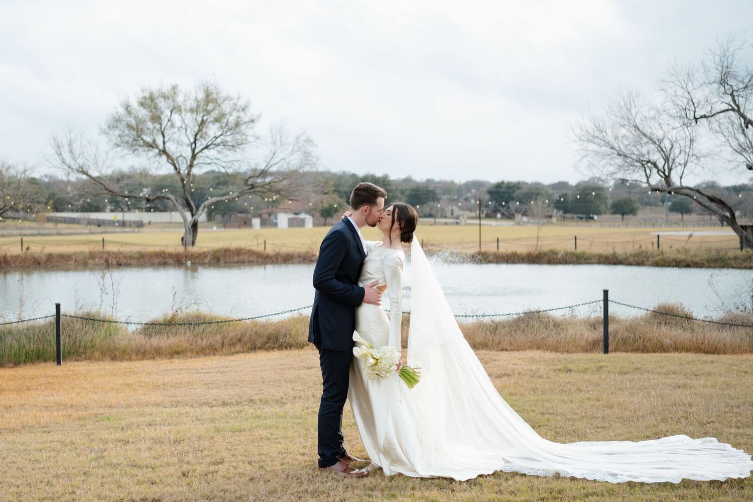 Wedding couple walking through an open field with bare trees and a long white dress train flowing behind them.