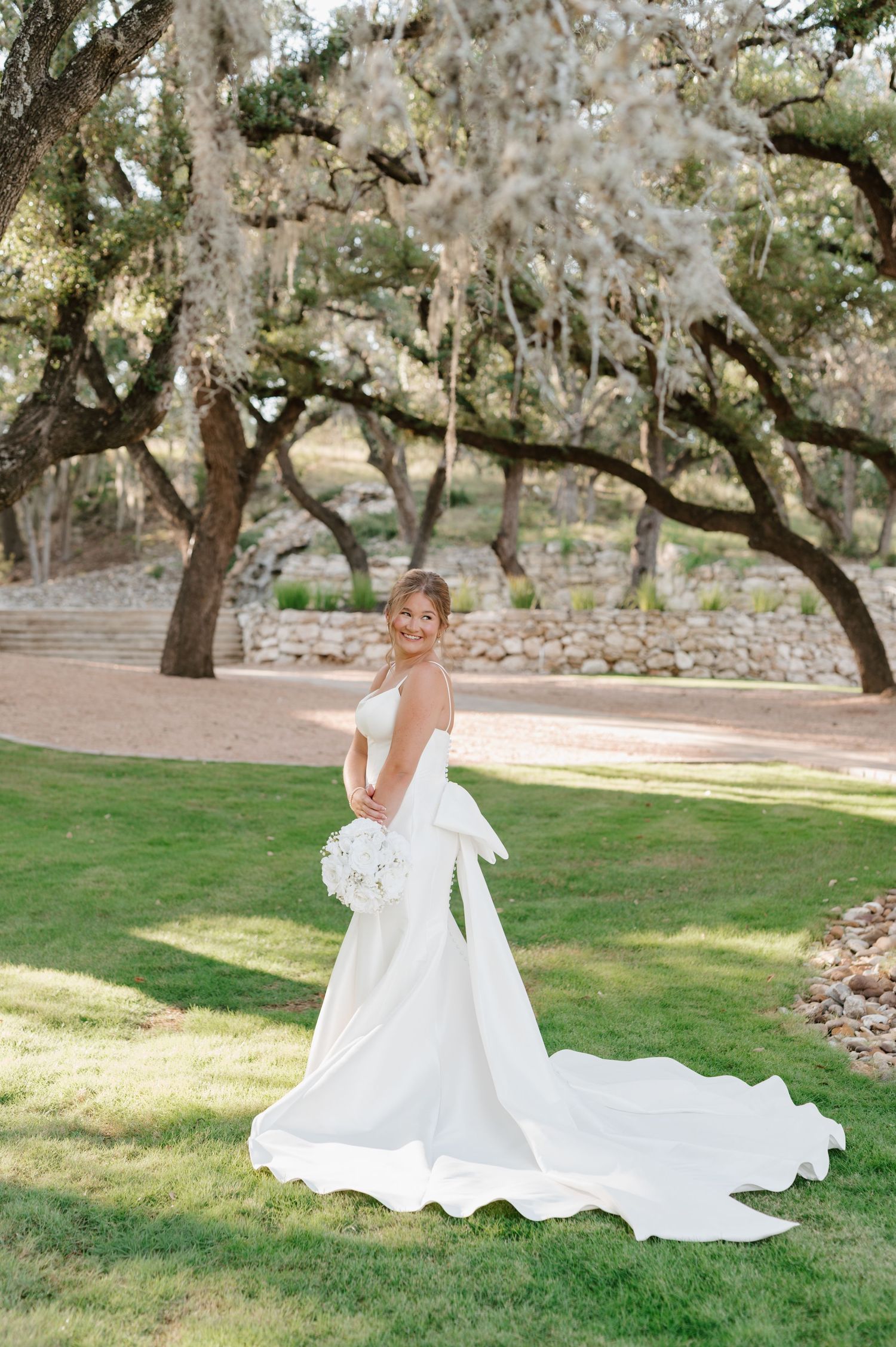 A white wedding dress flows across green grass under moss-draped oak trees in a romantic garden setting.