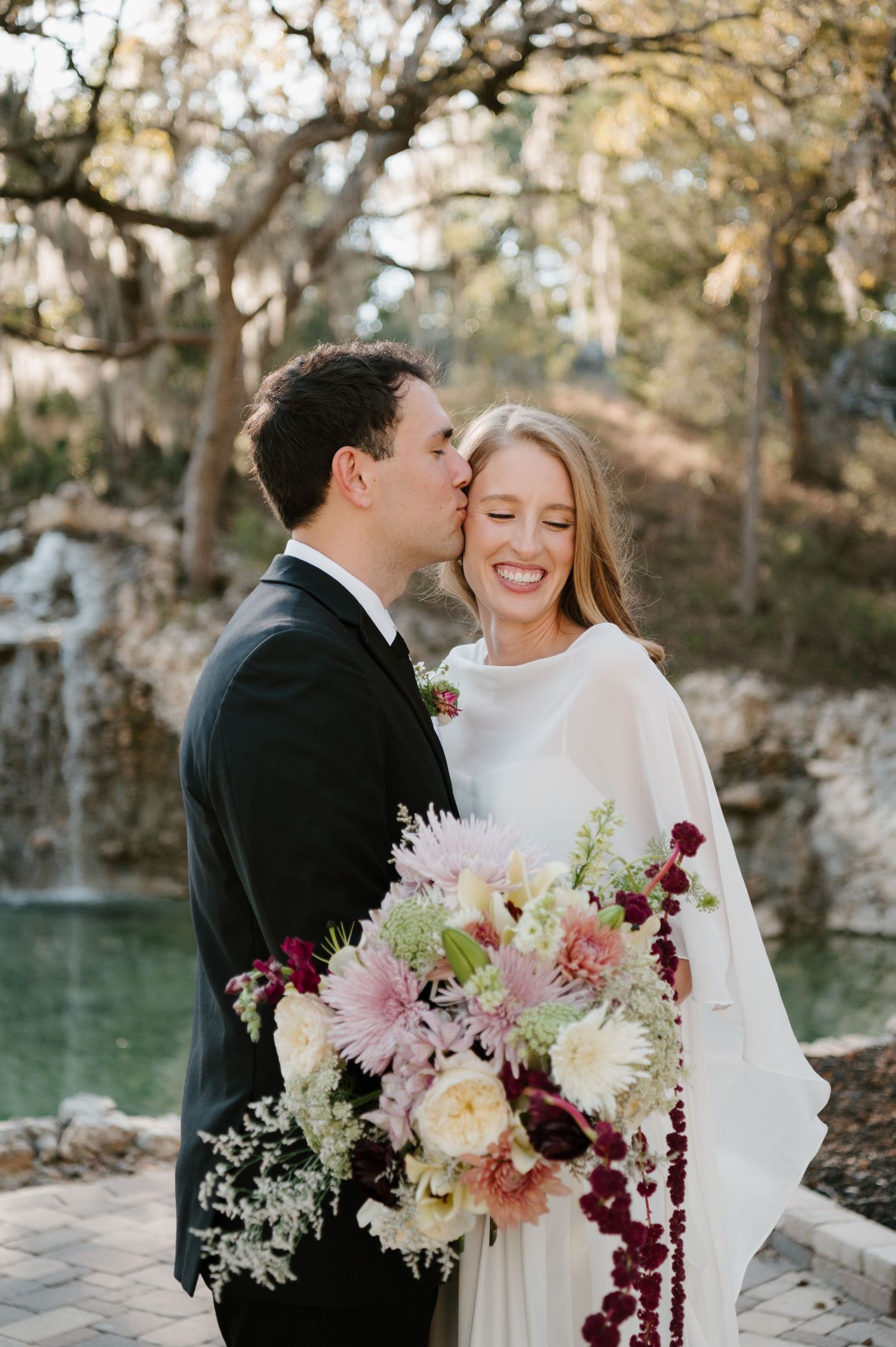 Wedding couple embracing outdoors with pink and burgundy floral bouquet.