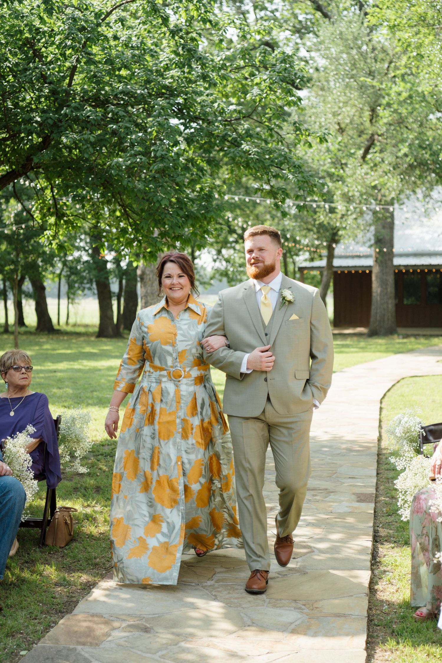 A groom in a sage suit escorts a woman in a floral yellow and blue dress down an outdoor wedding aisle.