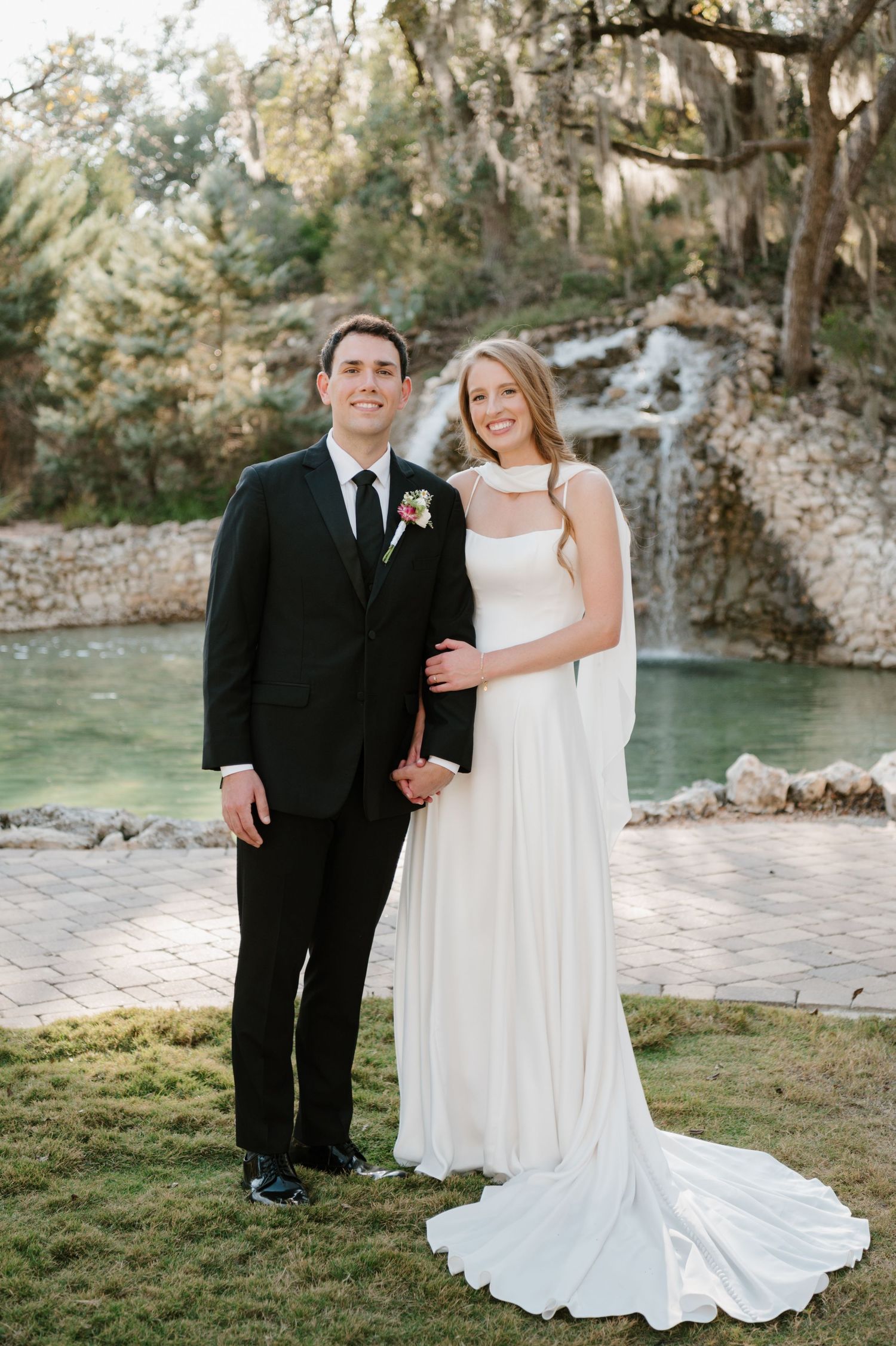 Elegant wedding couple poses near water with stone walls and trees in background.
