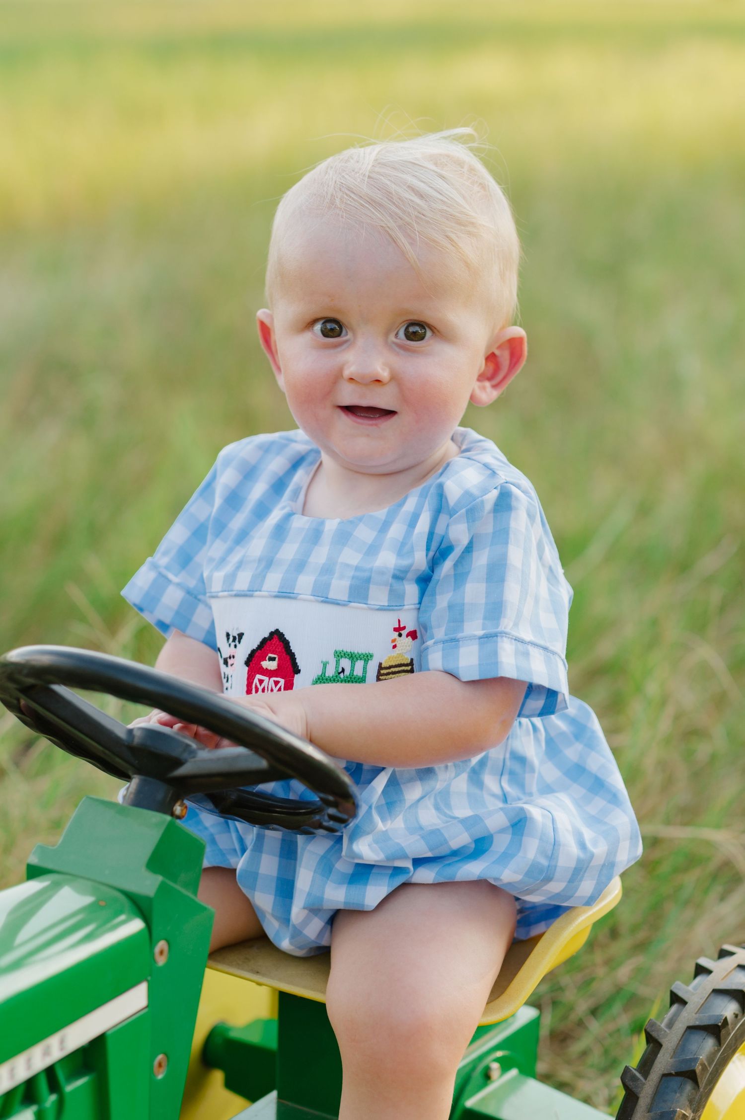 A baby in blue gingham sits on a green toy tractor in a sunny outdoor setting.