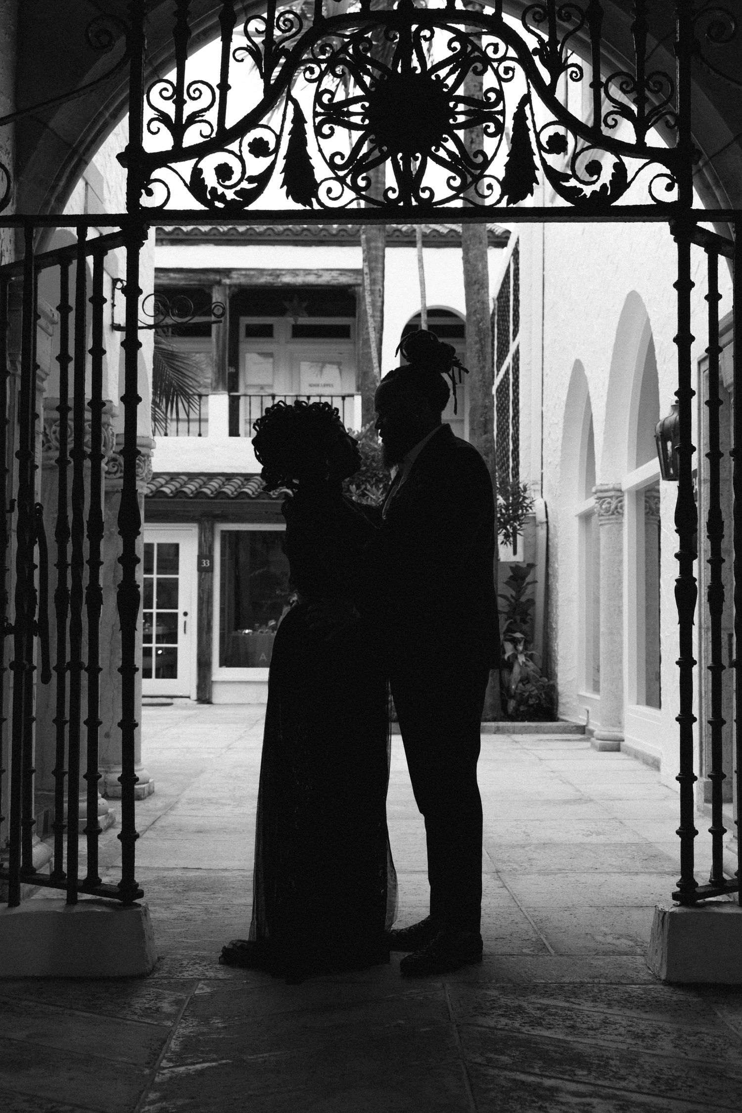 A silhouetted couple stands beneath an ornate wrought iron archway in a black and white photograph.