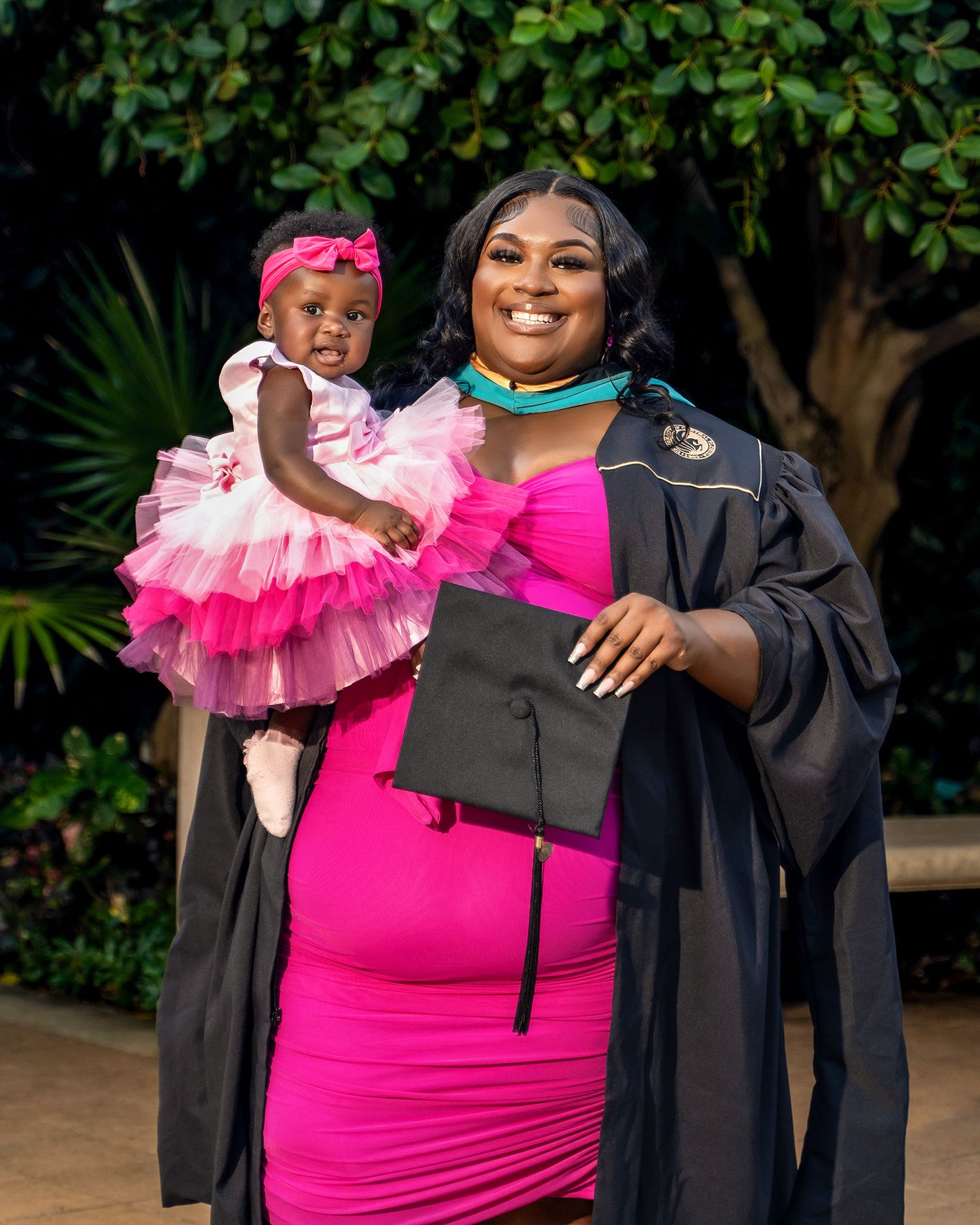 Graduate in academic regalia poses with small child wearing pink tulle dress outdoors.