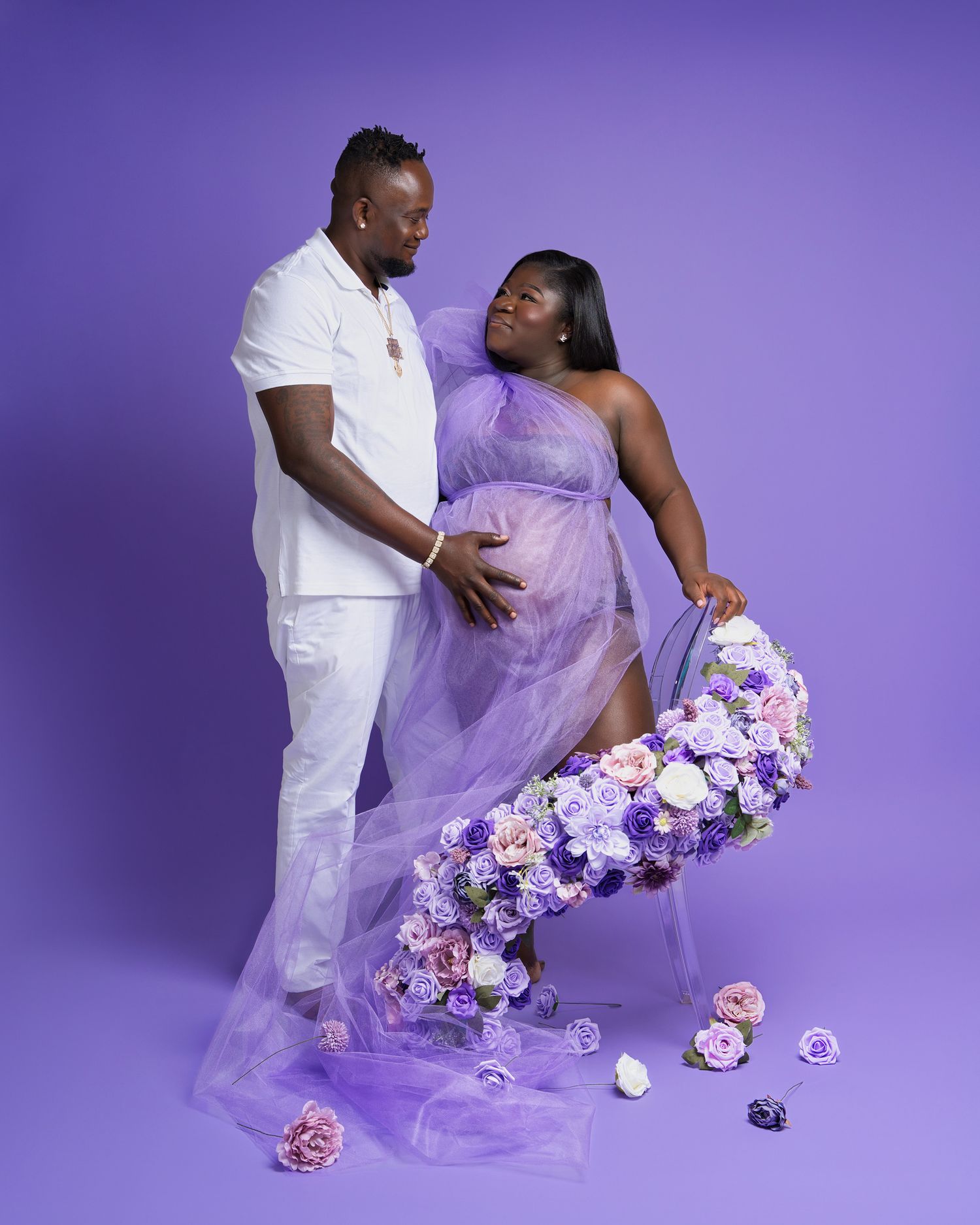 Romantic maternity portrait with purple dress and floral decorations against purple studio background.