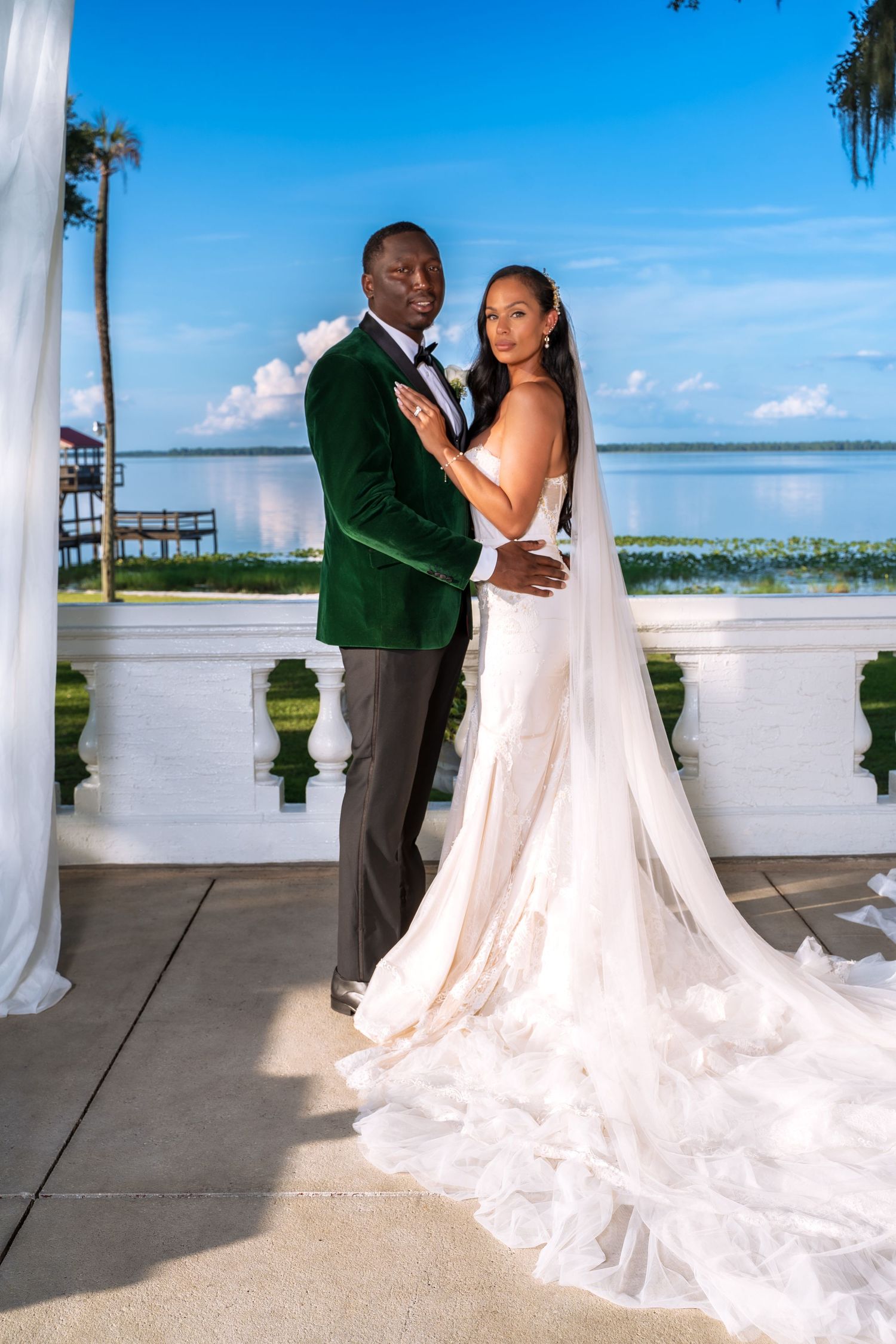 A bride in white gown and groom in emerald velvet jacket share an intimate moment at waterfront venue.