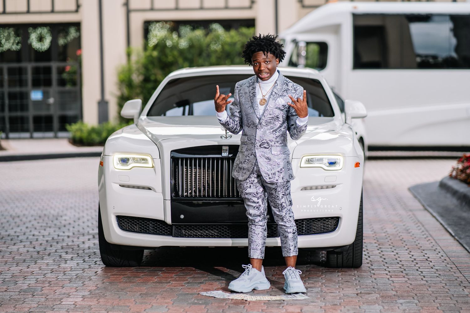Someone in a silver patterned suit poses in front of a white Rolls Royce.