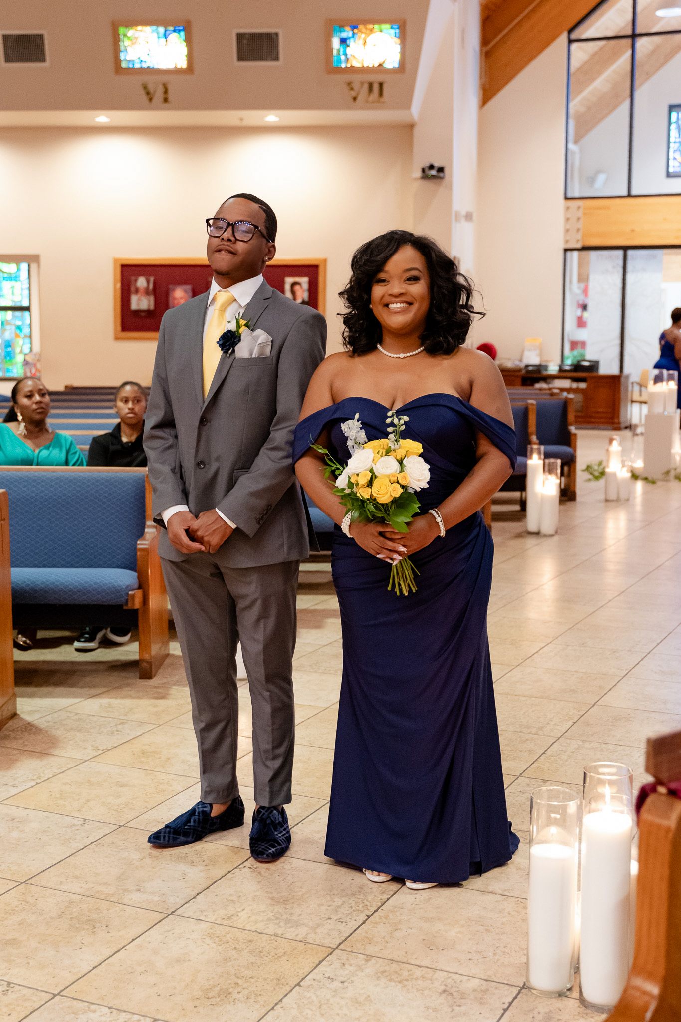 Wedding party member in grey suit stands with bridesmaid in elegant navy blue dress holding yellow bouquet.